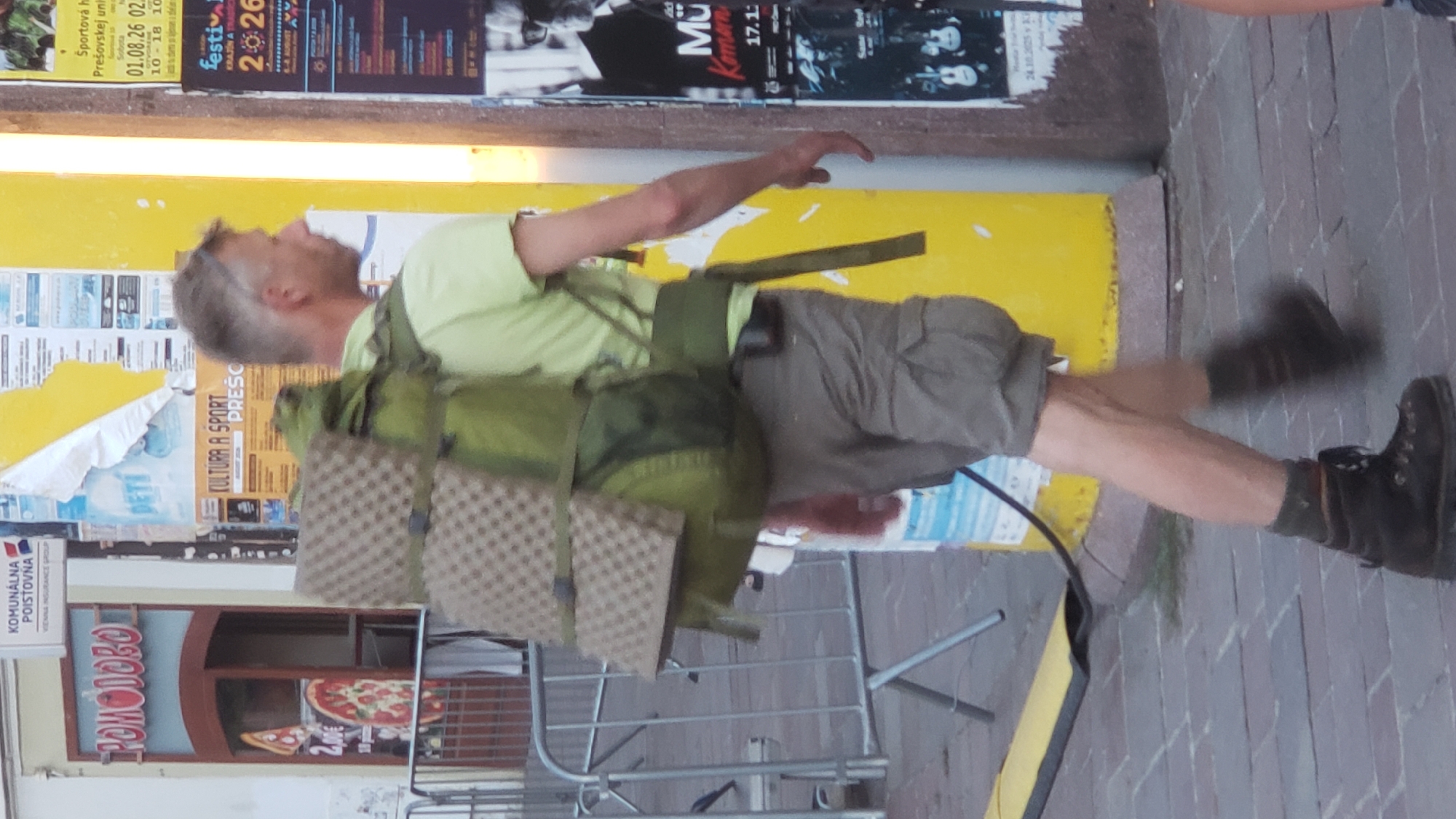 A person with a large backpack walks down a sidewalk in the city of Prešov, Slovakia. The person is wearing a light green t-shirt, khaki shorts, and dark hiking boots. The backpack is green and has two padded sleeping pads attached to the top. The background shows posters on a yellow pillar, and a building with signage that includes the word "POMODORO". The sidewalk is paved with bricks.