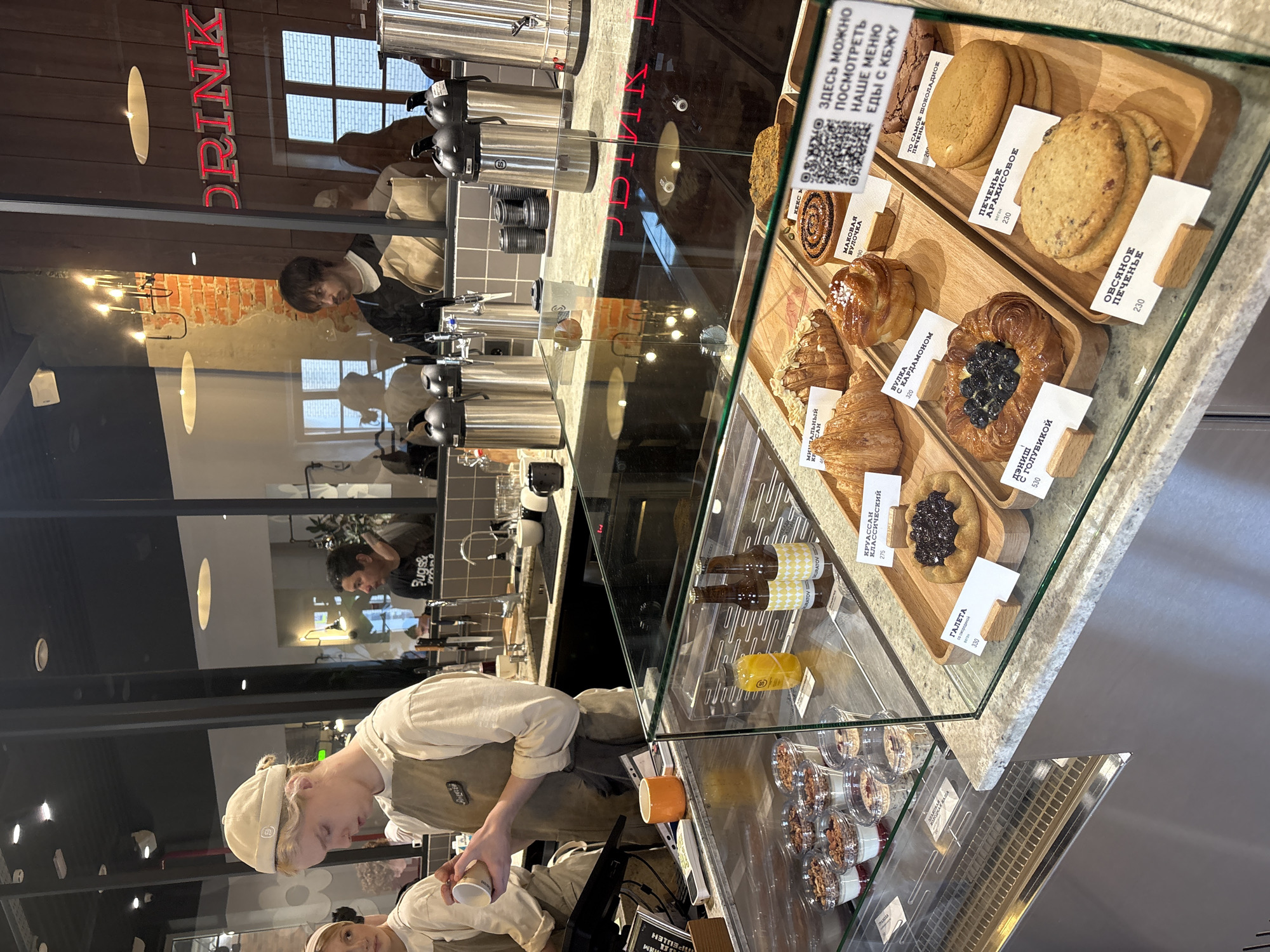 Here is a detailed description of the image:
**General Description:**
The image appears to be taken inside a bakery or cafe, possibly in Moscow, Russia, based on the signage. The scene showcases a display of pastries and other food items, along with staff members working behind the counter.
**People:**
* Two people, likely staff members, are visible. One is in the foreground, wearing a beige hat and apron, possibly preparing a drink. Another person is partially visible.
* Reflections in the glass show additional individuals, including one person wearing a dark shirt with the word "Bugs&" on it.
**Objects:**
* **Display Case:** A glass display case holds a variety of pastries, including cakes, cookies (like peanut and oatmeal cookies), danishes, and croissants. Each item has a label with its name and price in Russian rubles.
* **Food Items:** Cakes, buns, rolls, pastries and cookies
* **Drinks:** Juice, water, and other bottled drinks
* **Coffee/Tea Station:** A stainless steel coffee or tea station with multiple dispensers is visible behind the counter.
* **Signage:** There is a red neon sign that reads "DRINK". Another sign with a QR code invites customers to view the menu.
**Actions and Interactions:**
* One staff member is actively preparing a drink.
* Customers might be looking at the pastry selection.
**Environment:**
* **Interior:** The interior has a modern aesthetic with tile walls.
* **Lighting:** The space is well-lit with a mix of natural and artificial light.
* **Cleanliness:** The bakery appears clean and well-organized.
**Location Clues:**
* The Russian text on the food labels and signage indicates that the location is likely in Russia.
* Specific menu items and the style of the bakery could potentially suggest Moscow or another major Russian city.