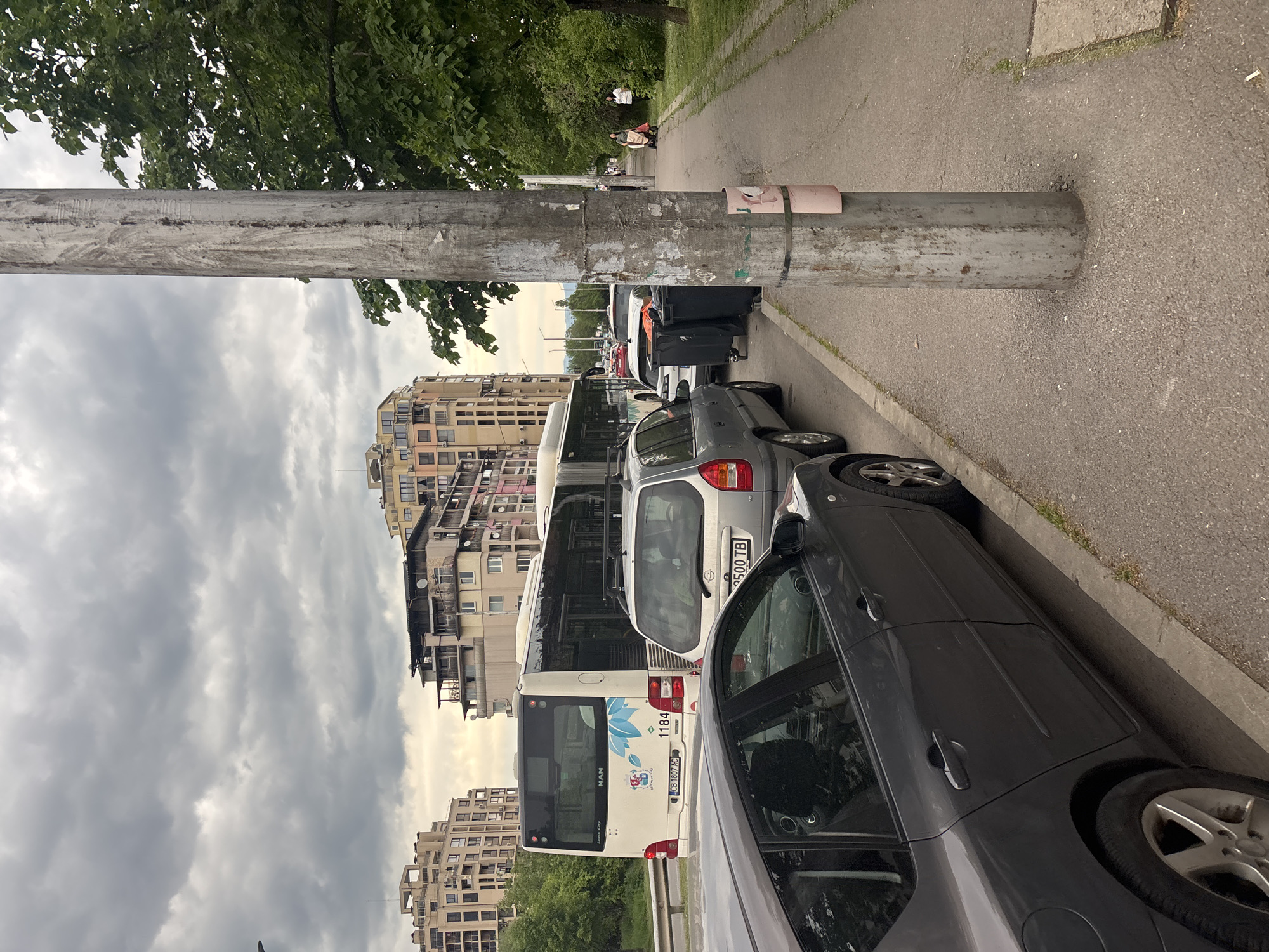 The scene depicts a street in Sofia, Bulgaria with parked vehicles along a sidewalk. A gray car is in the foreground, followed by a silver car and a white bus. Luggage is placed next to the cars. A metal pole is in the foreground obscuring the view. Buildings are in the background, and the sky is cloudy. Some people are visible walking on the sidewalk.