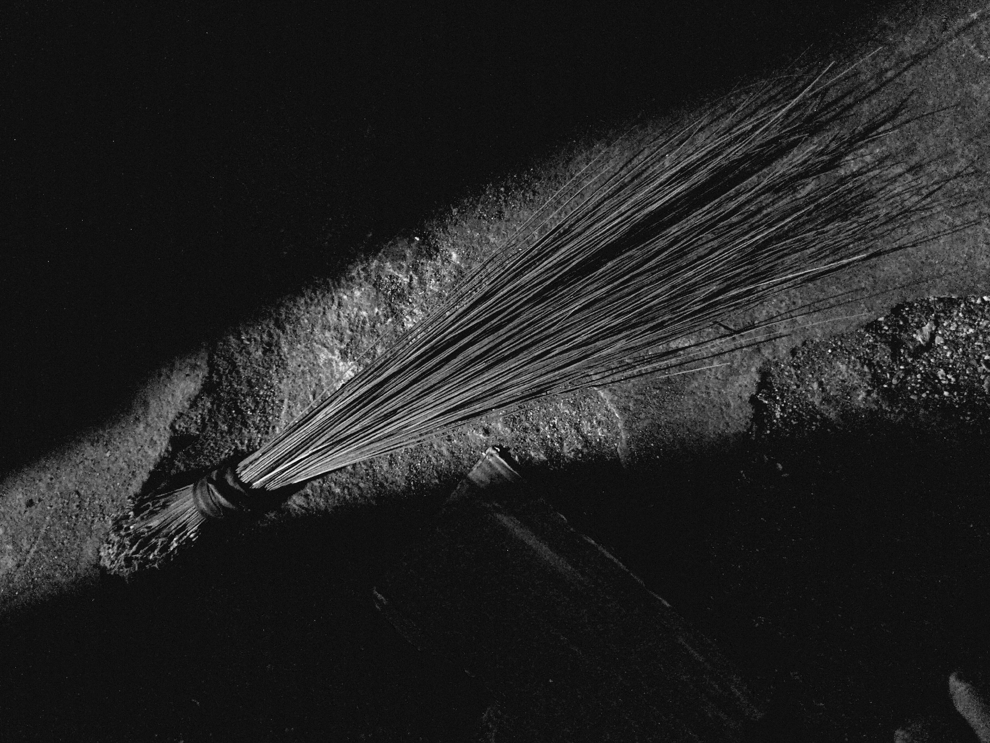 The black and white image depicts a traditional broom made of numerous thin, stiff bristles, resting diagonally across a rough, textured ground surface. The broom's handle end, where the bristles are tightly bound, is positioned towards the bottom left, while the sweeping end fans out towards the top right.
A strong, directional light source illuminates the broom and a band of the ground beneath it, creating sharp highlights and deep, contrasting shadows. This chiaroscuro lighting effect emphasizes the individual bristles of the broom and the uneven, granular texture of the ground, which appears to be concrete or compacted dirt. Much of the foreground and background beyond the light strip is plunged into almost complete darkness, intensifying the focus on the broom.
No discernible activity is taking place; the broom appears to be left momentarily on the ground. The stark lighting and monochrome palette highlight the utilitarian object as a subject of abstract form and texture. There is no visible text in the image. The precise location or time of day is not indicated, but the intense, directional light suggests either direct sunlight in a partially shaded area or a focused artificial light source.