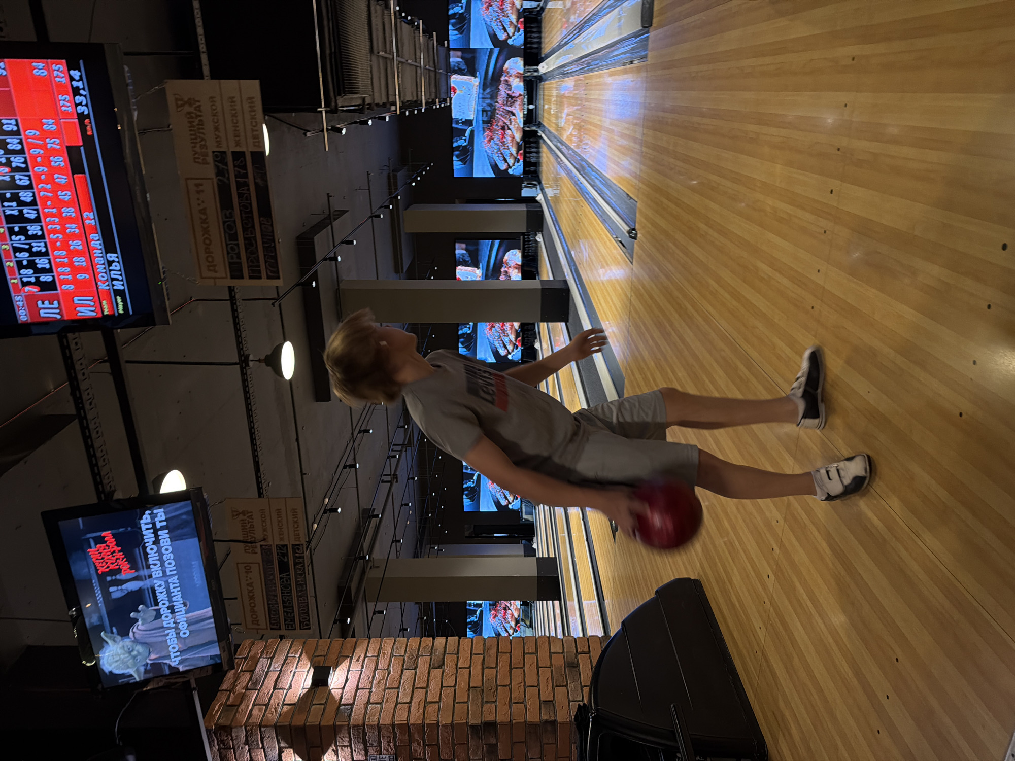 This media file depicts a young person bowling in an indoor bowling alley. The person, with blond hair, is wearing a gray t-shirt and shorts and is in the process of throwing a red bowling ball. The scene is set in a bowling alley with polished wooden lanes stretching into the distance. Large screens are visible in the background displaying graphics, and overhead lights illuminate the area. The overall atmosphere appears to be casual and recreational.
Several pieces of text are visible in the image. On a screen to the left, a scoreboard displays scores and player names, with some text in Russian, including "Команда 12" (Team 12) and "ИЛЬЯ" (Ilya). Another screen shows a still image and Russian text which translates to "To turn on the lane, call the waiter." Signs above the lanes also have Russian text, indicating lane numbers, results, and categories like "МУЖСКОЙ" (Male), "ЖЕНСКИЙ" (Female), and "ДЕТСКИЙ" (Children). The time "00:45" is also displayed on the scoreboard. The setting suggests an evening or late-night activity, given the lighting and general ambiance. The person's posture and focus on the ball suggest engagement in the activity.