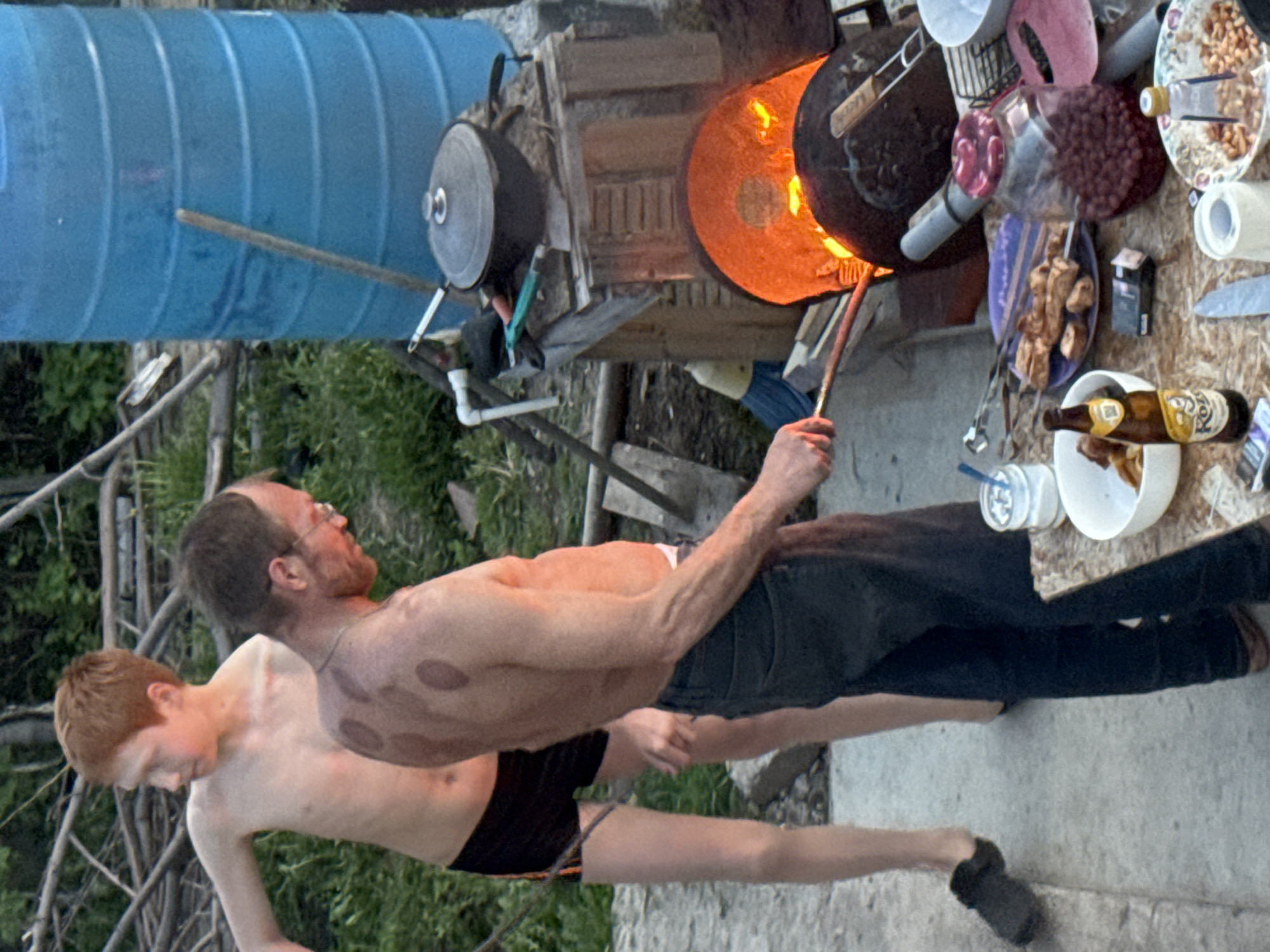 Two shirtless men are standing on a concrete surface. One is holding a long metal tool near an open fire with a glowing orange interior. On the right side of the image, there's a makeshift table with various objects on it, including plates of food, containers, and a beer bottle. In the background, there's a large blue container, branches, and some greenery. The location is Chaikovsky, Russia.