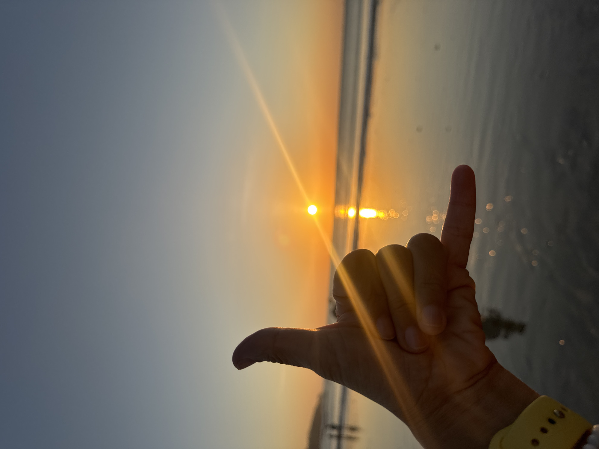 A person's hand in the foreground forms a "shaka" sign with their thumb and pinky finger extended, against a backdrop of a beach at sunset. The sun is setting over the ocean, casting a warm orange glow on the sky and reflecting on the wet sand. The waves are gently rolling in, and the water is calm. In the distance, a few blurred figures can be seen walking along the shoreline. The image appears to be taken in Kuta Selatan, Indonesia, a popular tourist destination known for its beautiful beaches and sunsets.