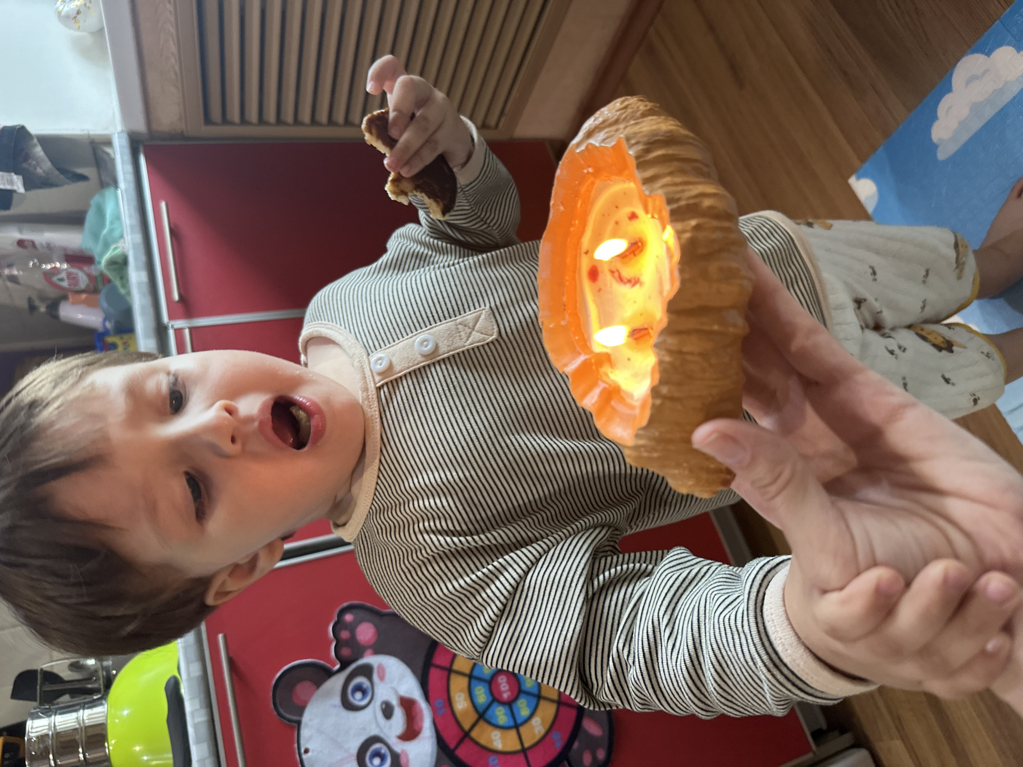 In this close-up, eye-level shot, a young child with brown hair and open mouth is holding a decorative pumpkin-shaped candle with two lit wicks. The child is wearing a horizontally striped long-sleeved shirt and patterned shorts. In their right hand, they are holding a piece of food to their mouth. Another hand, presumably an adult's, is supporting the candle from below. In the background, a red cabinet is visible, along with a colorful dartboard featuring a panda graphic. The environment appears to be a domestic setting, possibly a kitchen, with wooden flooring and a blue mat with cloud patterns. The image's angle and composition suggest it was taken indoors. No specific details point to the city of Vladivostok, Russia.
