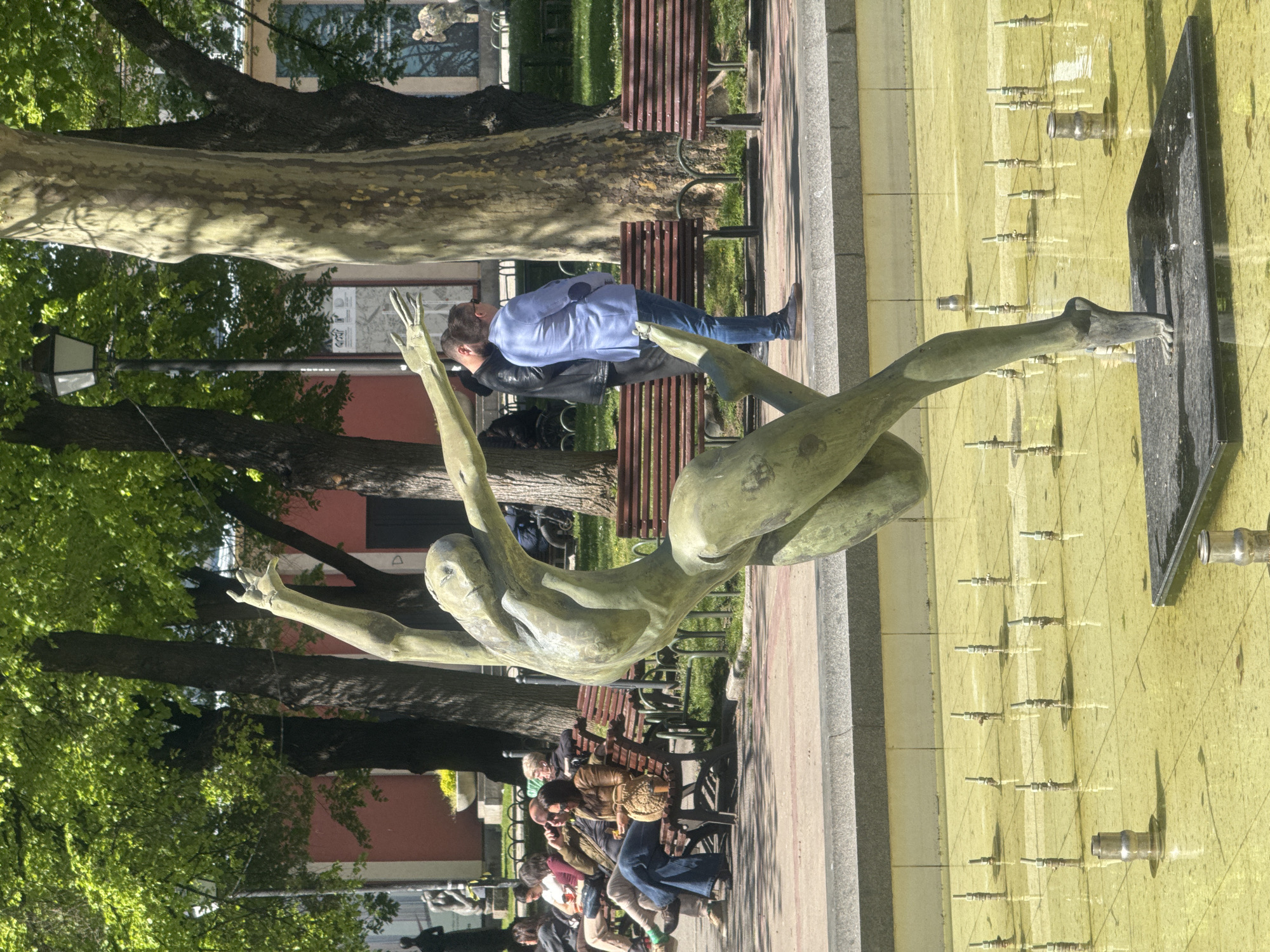 The image shows a bronze statue of a person stretched out horizontally, presumably a diver or swimmer, positioned above a fountain in a public space. The fountain has a rectangular basin with multiple vertical jets of water. A sidewalk runs parallel to the fountain. People are sitting on benches alongside the walkway. One man is standing near the statue looking at it. Trees are visible in the background. The location is in Sofia, Bulgaria.
