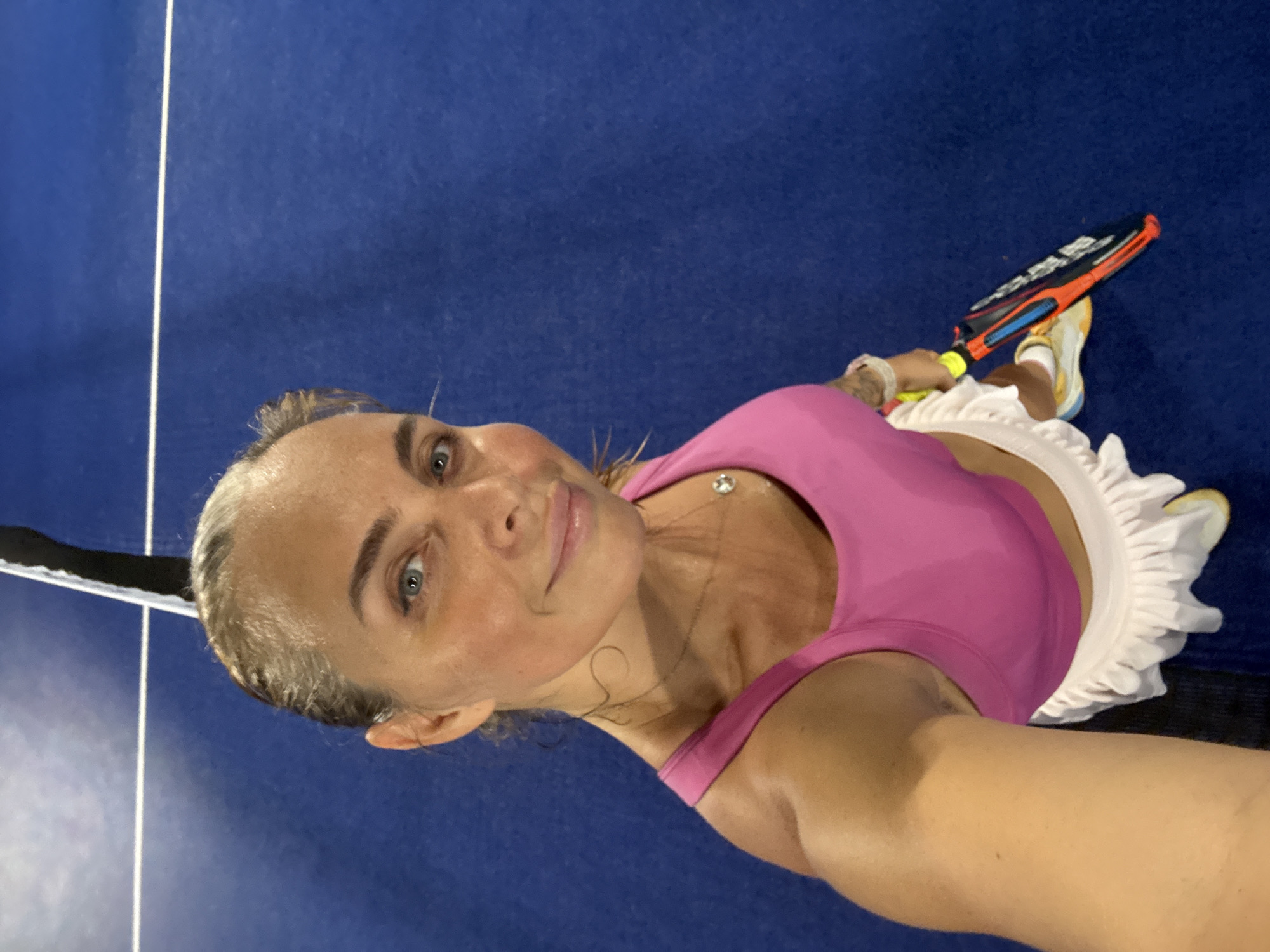 This is a media file that shows a woman on a blue sports court, possibly a tennis or padel court, as indicated by the net visible in the foreground and the racket she is holding. The woman is wearing a pink sports bra and a white ruffled skirt. She has light-colored hair and appears to be looking at the camera with a smile. Her skin appears slightly flushed, suggesting she may have been playing sports or exercising.
The setting is indoors or outdoors with artificial lighting, given the consistent illumination and the blue court surface. There is no indication of the time of day or weather conditions. The woman is holding a racket with orange and black accents. She is also wearing athletic shoes.
The image is taken from a high angle, looking down at the woman. The focus is on the woman, but the court and some sports equipment are also visible. The woman's expression suggests a positive or content mood. There is a faint necklace with a pendant visible around her neck. The overall scene depicts a moment of leisure or sports activity.