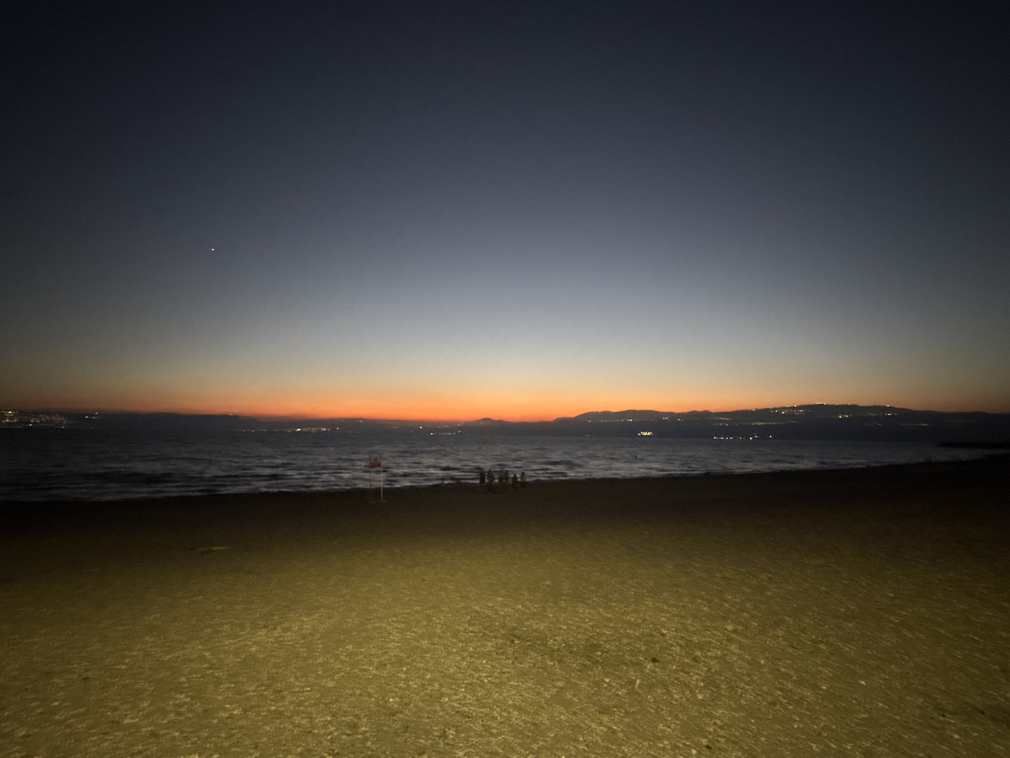 The image depicts a twilight scene at a beach, possibly near the Sea of Galilee, within the Golan Regional Council, Israel. The sky transitions from a deep navy at the top to a gradient of lighter blues and finally to a vibrant orange and red band just above the horizon, indicating the last moments of sunset or the first hints of sunrise. A single bright star is visible in the upper left quadrant of the sky.
The sea is visible in the middle ground, with gentle waves lapping at the shore. The water appears dark due to the low light conditions. Along the distant shoreline, faint lights suggest the presence of settlements or a city. The silhouette of hills or mountains is visible in the far distance.
In the foreground, the beach is covered in sand and small pebbles. A small group of people, appearing as dark silhouettes, are gathered near the water's edge. A red and white lifeguard pole or marker is present on the beach, to the left of the group. The lighting on the sand suggests it is illuminated by an artificial light source, possibly a spotlight or streetlamp, creating a warm, yellowish glow on the ground. The overall atmosphere is calm and serene, capturing the transition from day to night.
