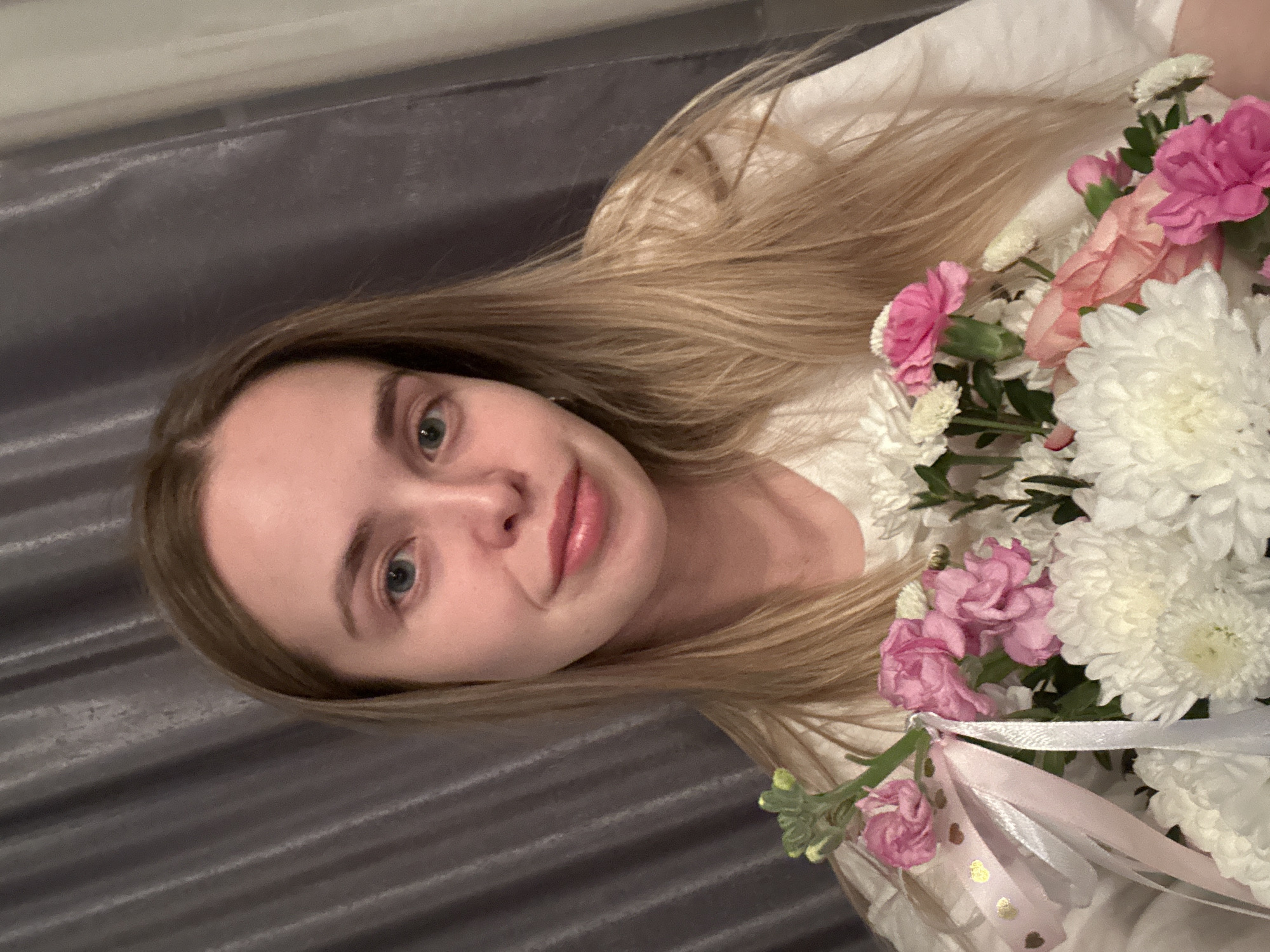 A woman with long, light-brown hair is holding a bouquet of pink and white flowers. The woman is fair-skinned with green eyes and pink lips. She is wearing a white shirt. The background is a gray, draped material. No definitive evidence within the image confirms the location as Chaikovsky, Russia.