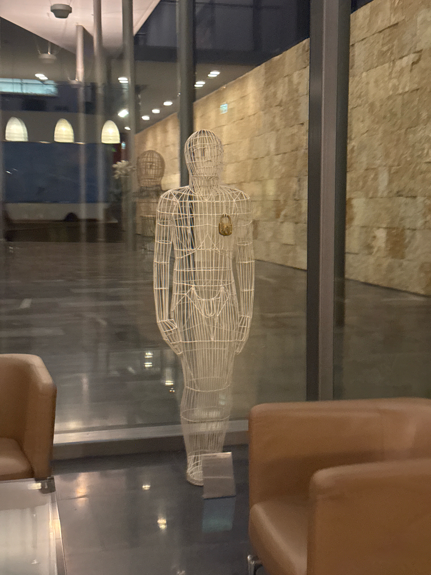 The image shows an interior scene, likely a modern building lobby or lounge, with a wireframe sculpture of a human figure as the central focus. The sculpture is white and has a small, gold-colored lock hanging from its chest. Behind the sculpture, a wall constructed of large, textured beige stones is visible. The floor is dark and reflective, with some light sources appearing as reflections.
In the foreground, partially visible on the left and right sides of the frame, are the arms and backrests of two modern lounge chairs, upholstered in a light brown or tan material. A portion of a glass-topped coffee table with a metallic frame is visible in the lower left corner.
The background features large glass windows or walls, offering a view of an adjacent space. Through the glass, architectural elements like ceiling lights, arched window shapes, and possibly other sculptures or furniture can be discerned. The lighting in the scene is subdued, with a mix of ambient light and artificial illumination creating soft highlights and shadows.
While the image does not contain explicit indicators of its specific location within Strovolos, Cyprus, the architectural style and material choices suggest a contemporary commercial or public building. The presence of modern furniture and a prominent art installation is consistent with such a setting.