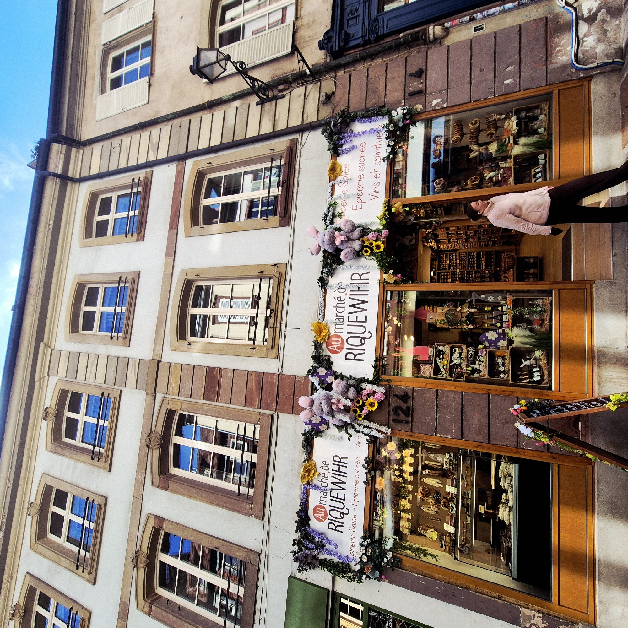 The image shows a building in Strasbourg, France, with a shop front decorated with flowers and signs that read "Au marché de RIQUEWIHR" and "Epicerie Salée - Epicerie sucrée - Vins et spiritueux." A woman is looking into the shop window. The building has multiple windows with a light-colored facade. The shop's address, "124", is visible next to the window. There is a chalkboard outside the shop.