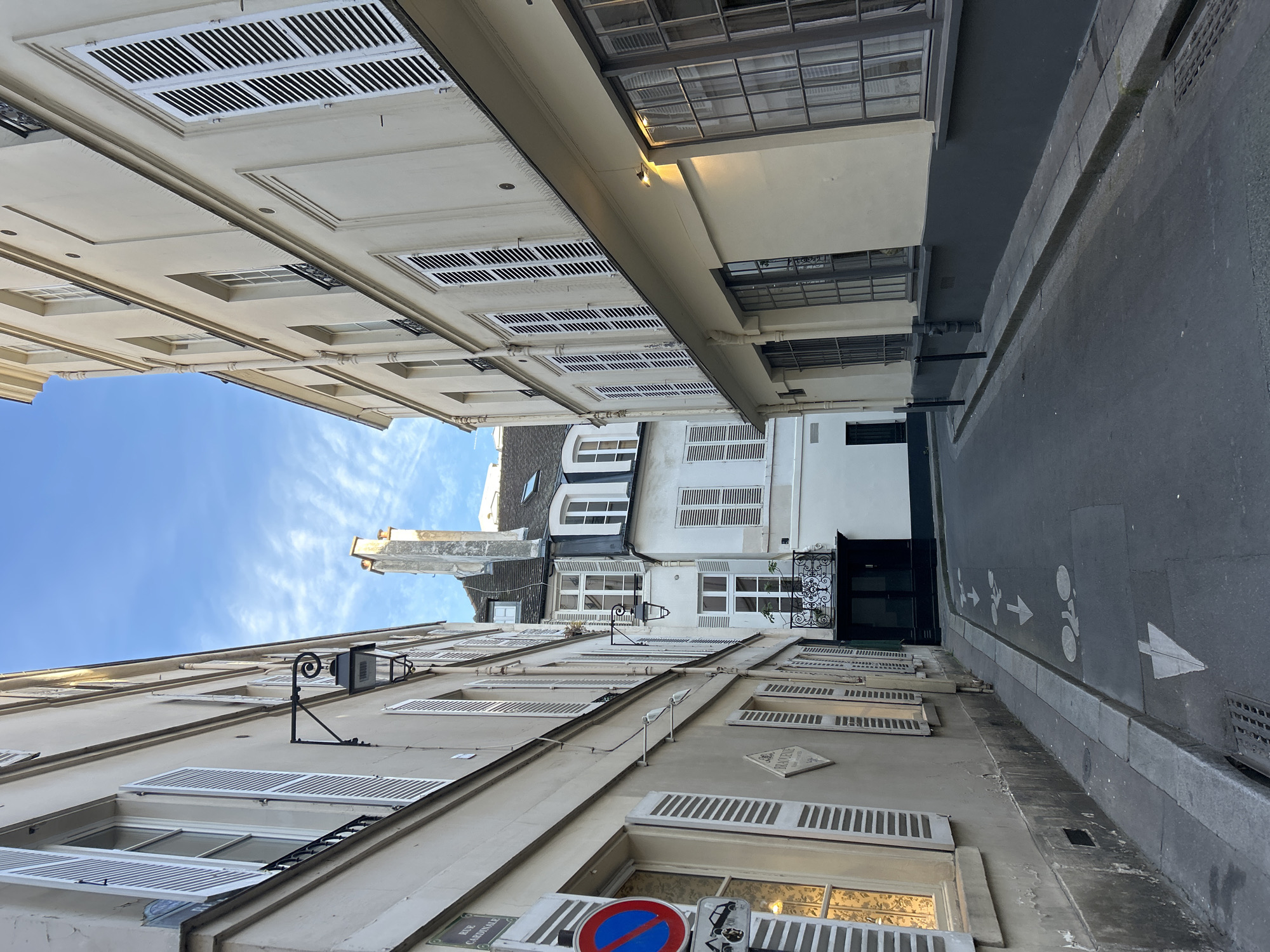 This image captures a narrow street in what appears to be an old European city, possibly Paris due to the architectural style of the buildings. The photograph is taken from a low angle, looking up along the street, which exaggerates the height of the buildings and the depth of the sky. The buildings on either side are multi-story structures with light-colored facades and numerous windows, many of which have white shutters. The shutters are a prominent feature, adding a repetitive pattern to the buildings' appearance. Several windows show glimpses of interiors. One building has a black awning or entranceway at the end of the street. Street lights, mounted on the sides of the buildings, are visible. Road signs are attached to one of the buildings, including what appears to be a no-parking sign. The sky is blue with thin, wispy clouds visible in the upper left corner of the image. The street itself is paved and has lane markings indicating a shared lane for bicycles and pedestrians. The overall impression is one of a quiet, historic urban environment. There are no people visible in the shot. The location is the city of Unknown, Unknown.