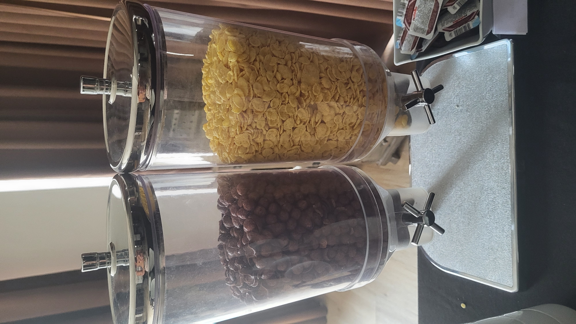 This image depicts a close-up, eye-level view of a hotel breakfast buffet setup, likely within a hotel in Prešov, Slovakia. The primary focus is on two transparent, cylindrical cereal dispensers with chrome lids and dispensing mechanisms. The left dispenser is filled with dark brown, spherical cereal, possibly cocoa puffs. The right dispenser contains light yellow, flake-shaped cereal, resembling corn flakes. Both dispensers are positioned side-by-side on a speckled silver tray with a metallic silver trim. The tray rests on a black surface, likely a tabletop or counter. Behind the dispensers, brown curtains create a soft, diffused background. To the right of the dispensers, a white rectangular container holds several individually packaged servings of what appears to be Nutella. No people are visible in the frame, and the scene is static, suggesting a prepared display for self-service. The lighting is soft, likely from ambient room light.