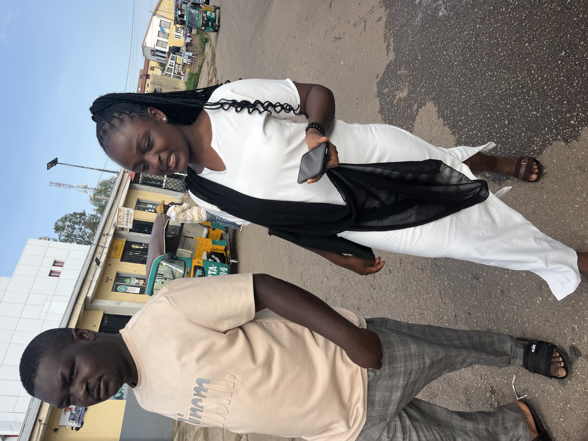 A medium shot captures a Black man and a Black woman walking on a paved road in Jos, Nigeria, under an overcast sky. The man, on the left, wears a light peach t-shirt with embossed lettering and gray plaid trousers, and black Nike slides. He is looking to his left, with a serious expression. The woman, on the right, wears a long white dress, a black scarf draped over her shoulders, and brown sandals. She holds a smartphone in her right hand and appears to be speaking, looking to her left. In the background, several buildings line the street, with a yellow-colored building featuring the inscription "P.I.O.S." prominently visible. Three-wheeled auto-rickshaws, commonly known as Keke Napep, are parked and in motion on the road. The scene depicts a typical street environment in Jos, with a blend of commercial structures and bustling street life.