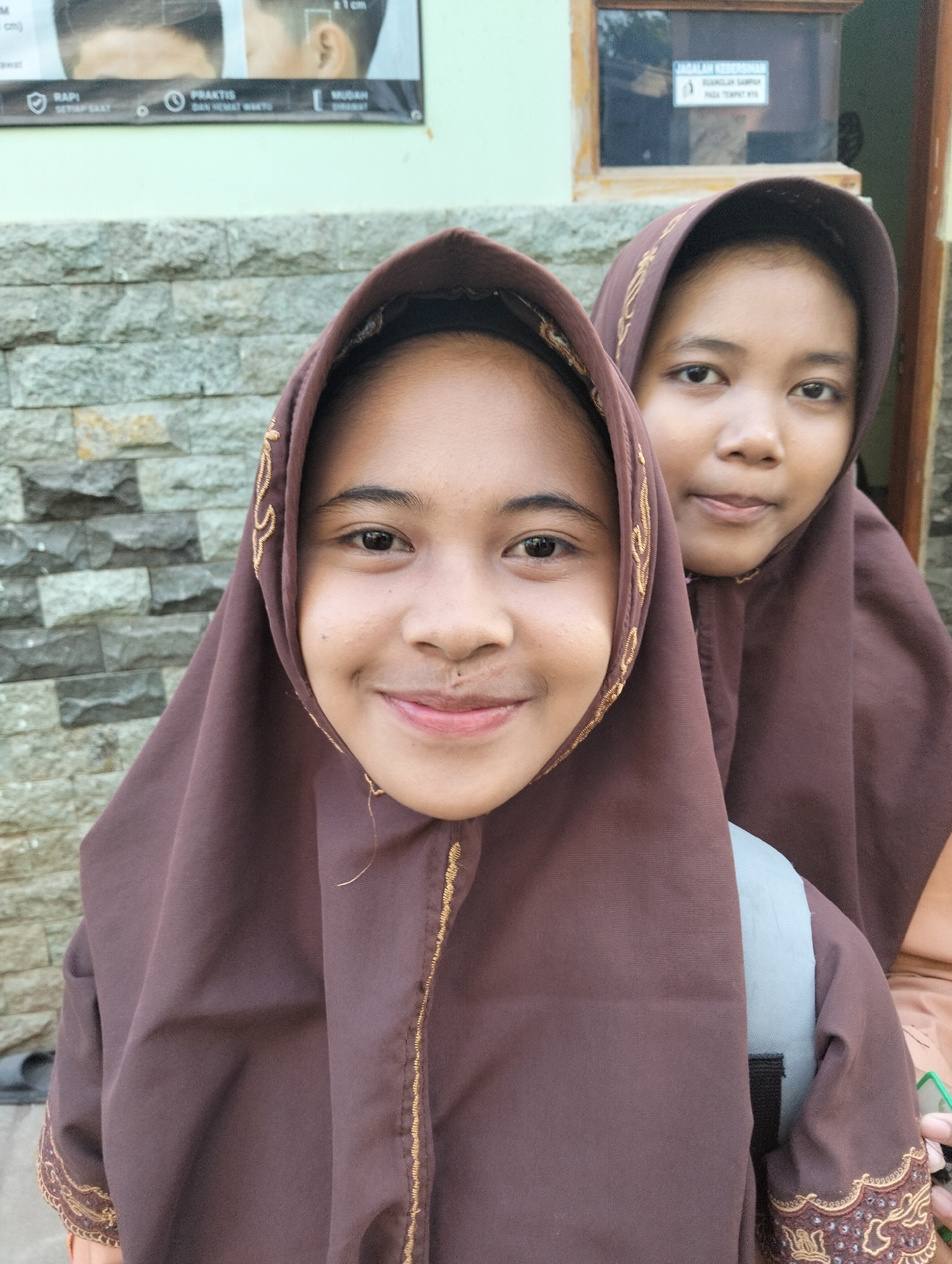 Two young girls wearing brown hijabs and school uniforms are posing for a photo outdoors. The girl in the foreground is closer to the camera and smiling, while the girl behind her is looking slightly away. They are standing in front of a stone wall. In the background, there is a building with a window and a sign. The scene appears to be in a public area, possibly a school or community center, given the presence of uniforms and the outdoor setting. The image was taken in Paciran, Indonesia.