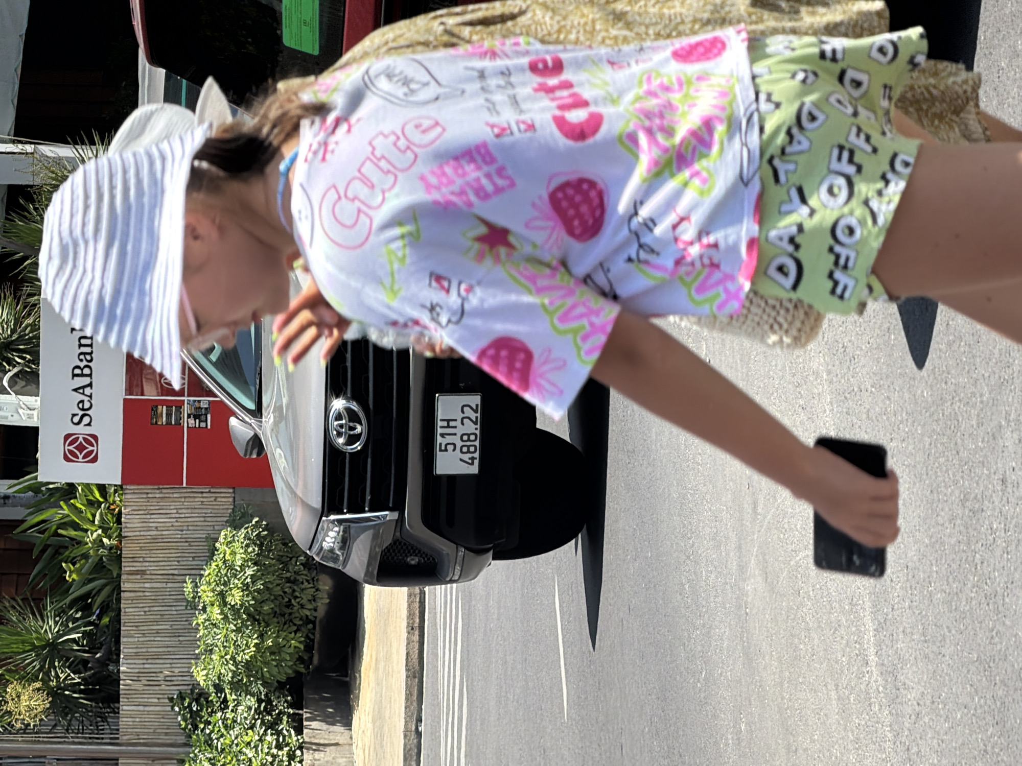 A young person wearing a white striped hat, glasses, and a white t-shirt with strawberry and text graphics walks across a street in Nha Trang, Vietnam. The t-shirt has the word "Cute" printed in pink multiple times, along with strawberries and other small graphics. The person is also wearing light green shorts with the words "DAY OFF" printed in white. They are holding a black smartphone in their left hand.
In the background, a grey Toyota Fortuner SUV is parked on the street. The license plate reads "51H 488.22". Behind the SUV is a building with a red facade and a bamboo-covered wall. A sign for "SeaBank" is visible on the building. Lush green bushes and plants are seen in front of the building and on the sidewalk. The street has white lane markings, indicating a crosswalk. The lighting suggests it is a sunny day.