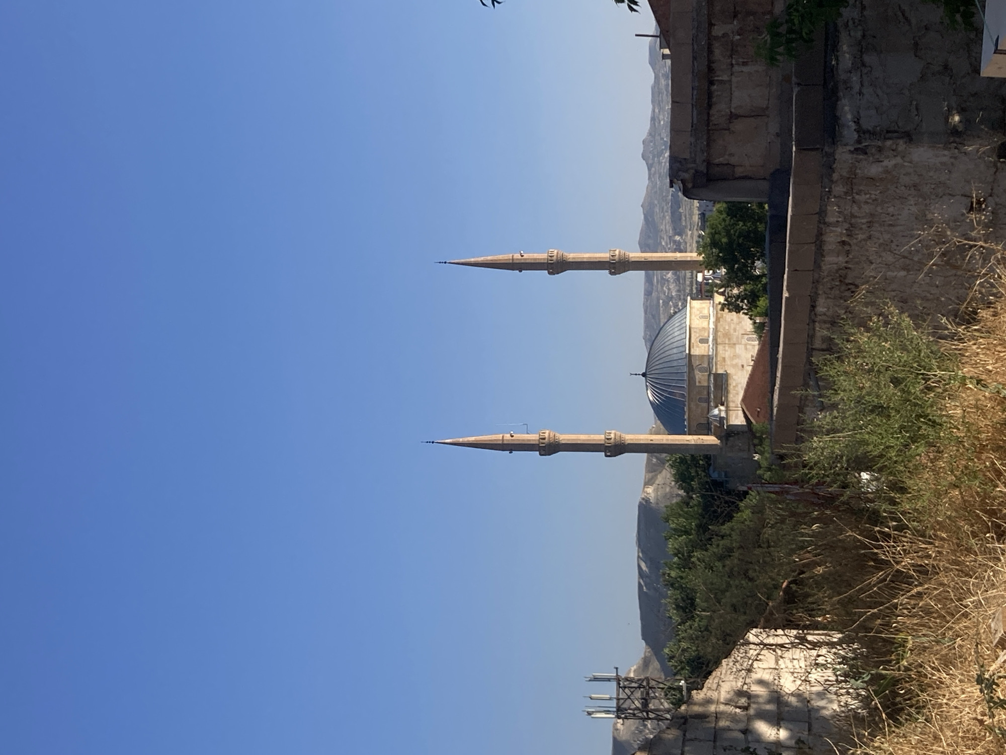 The image, taken in Avanos, Turkey, depicts a serene outdoor scene under a clear blue sky, suggesting a bright, sunny day. The orientation of the photo is rotated 90 degrees clockwise. In the mid-ground, a mosque with a prominent dome is visible, flanked by two tall, slender minarets. The minarets are intricately designed with multiple tiers and balconies. The architecture of the mosque and minarets points to a traditional Islamic style.
The foreground is dominated by a rough stone wall and dry, straw-like vegetation, hinting at a dry climate or season. Behind the mosque, a lush green tree adds a touch of life to the arid landscape, and further in the background, rolling hills or mountains can be seen, shrouded in a light haze, suggesting distance.
The scene appears to be a quiet, possibly religious site, with no immediate human activity visible in the main focus of the image, though a tiny figure can be seen on one of the minarets. The overall mood is peaceful and contemplative, amplified by the vast expanse of the sky and the timeless presence of the religious structure. The composition, with the mosque and minarets framed by natural and man-made elements, creates a sense of depth and perspective. There is no visible text in the image.
