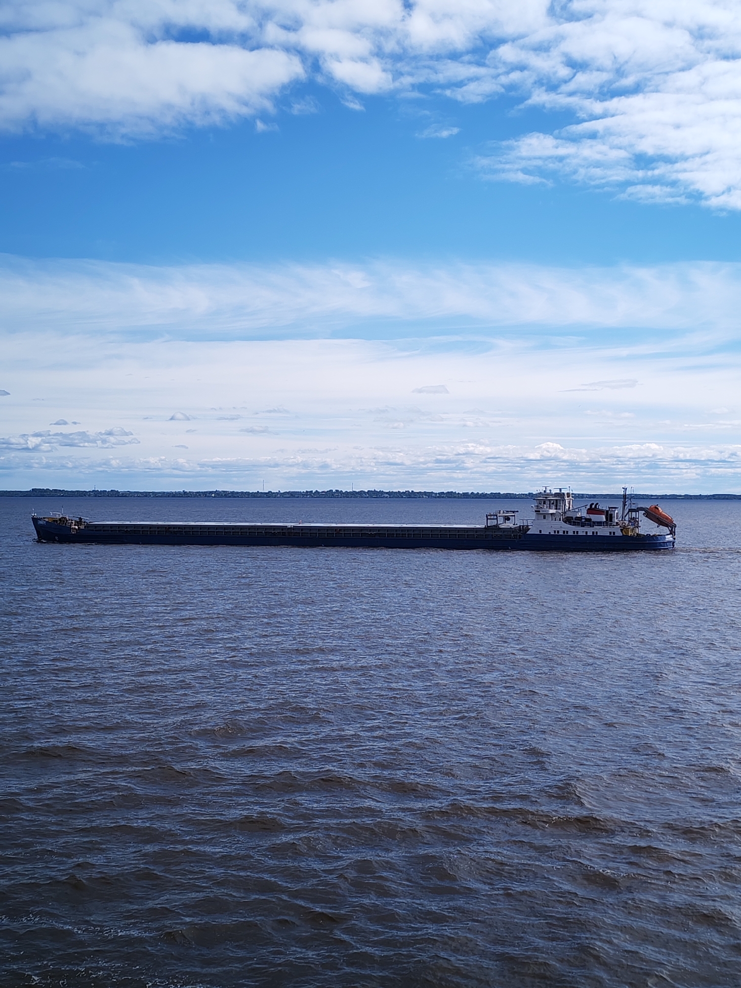 A barge traverses a body of water under a partially cloudy blue sky, the town of Belozersk, Russia, barely visible on the horizon. The barge is long and flat, likely carrying cargo, with a control structure at the far end. The water is dark and choppy, indicating a slight wind. There are no people visible in the scene.
