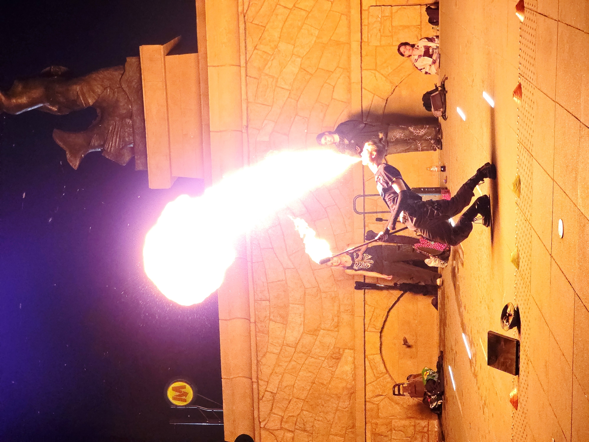 In Warsaw, Poland, at night, a performer is engaged in a fire-breathing act in front of a stone-paved plaza. The performer, dressed in dark clothing, is captured mid-action, expelling a large plume of flame from their mouth towards the left of the frame. Another performer stands to the right, also holding a flaming object. In the background, a stone wall with arched openings forms a backdrop, and a statue of a mermaid on a pedestal is visible at the top. A yellow "M" sign, indicative of the Warsaw Metro, is also present in the upper left corner. Several onlookers are seated or standing further back, observing the spectacle. The ground is illuminated by the fire and artificial lights, casting strong shadows.
