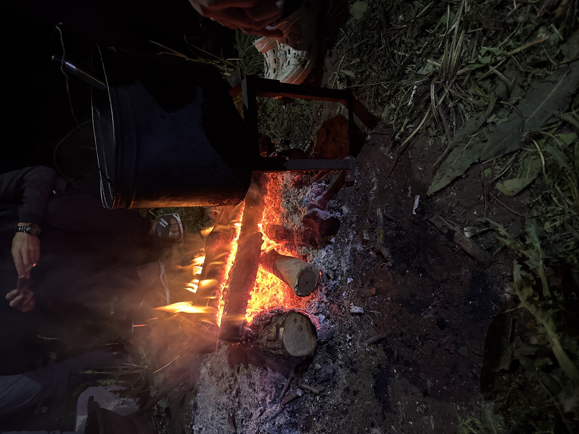 The image depicts a nighttime campfire scene likely in a rural or outdoor setting near Терси, Russia, based on the location request. A black pot hangs over a fire built with logs and kindling, with visible flames and embers. Ash and charred remnants surround the fire. Two people are partially visible in the image. One is wearing a dark jacket, with a hand and watch visible, holding what appears to be a small stick or match. Another person, wearing white shoes, is also visible near the pot and the fire pit. The surrounding environment includes dark vegetation, possibly grass and weeds. The scene appears to be set up for cooking or warmth around the campfire.