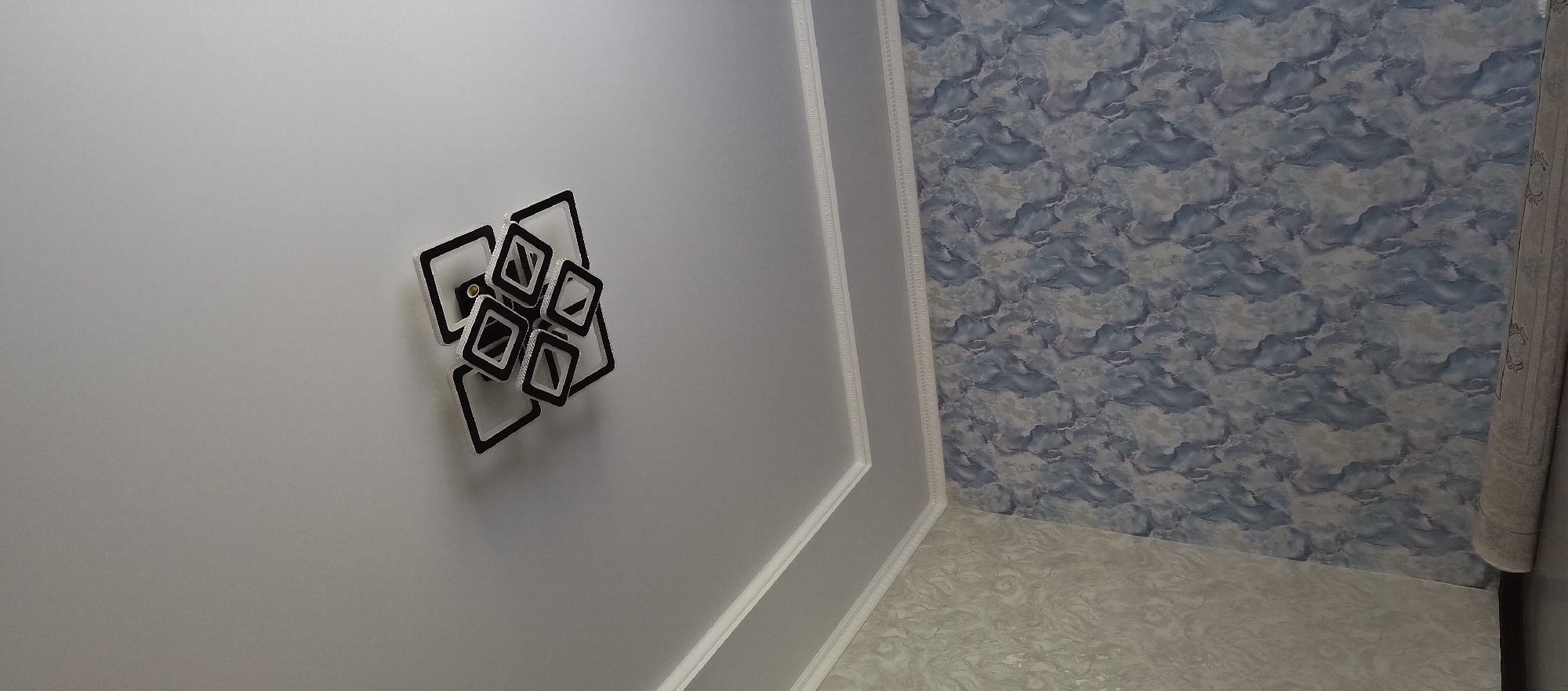 The image shows a modern, geometric ceiling light fixture with black and white square-shaped elements, suspended from a plain white ceiling. The ceiling has decorative white molding along the edges where it meets the walls. The walls are covered in wallpaper. The wall on the left has a light-colored, textured wallpaper with a marbled or abstract pattern. The wall on the right has a darker wallpaper with a blue and gray marbled or abstract pattern. A rolled-up carpet with a patterned edge is visible on the floor in the lower right corner of the image. The scene appears to be an interior of a room, likely a living room or dining area, in Barskoon, Kyrgyzstan. No people are present in the image.