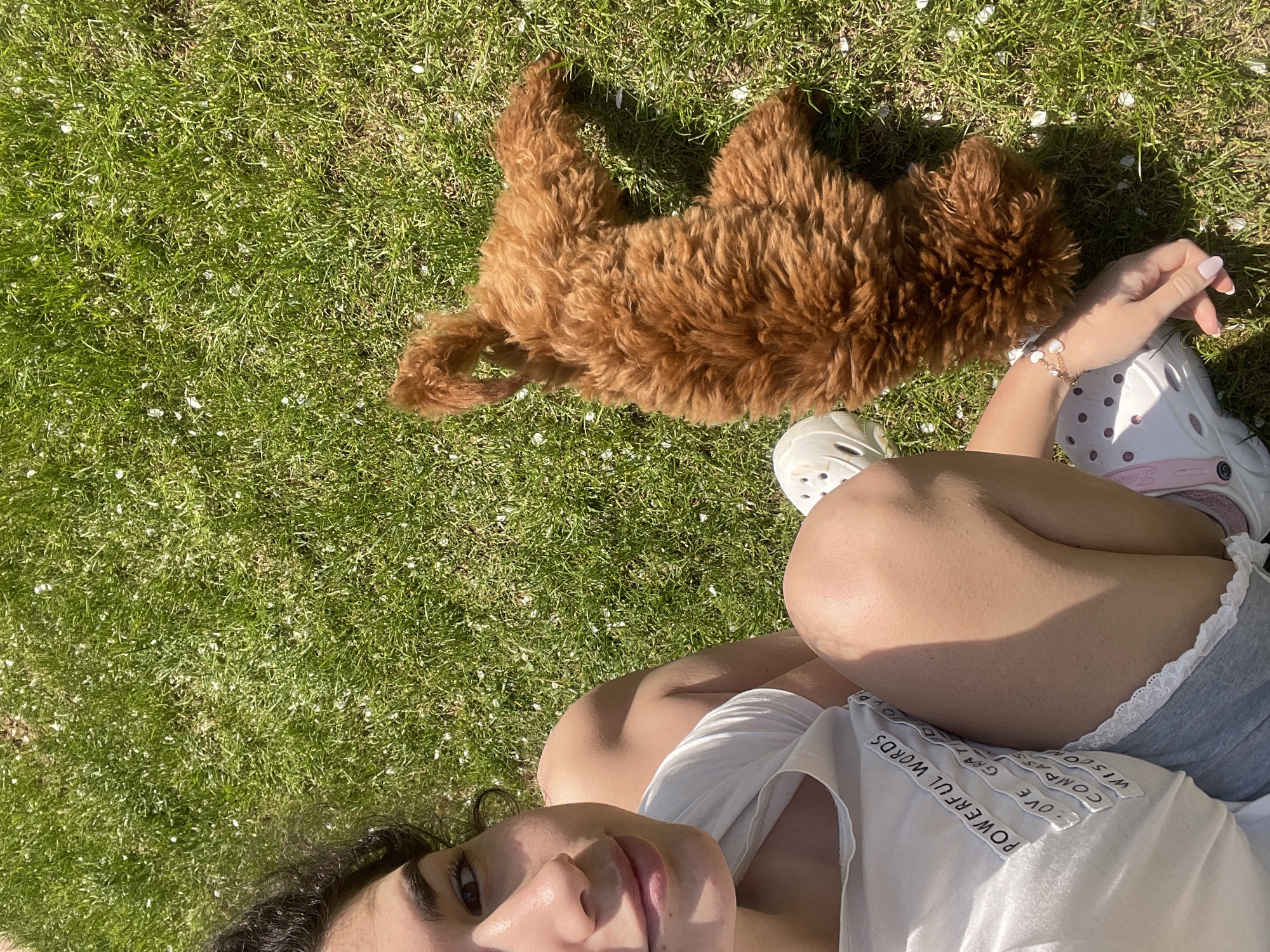 The image captures an overhead view of a person and a dog relaxing on lush green grass under bright sunshine. A female with dark curly hair is seen looking towards the camera with a gentle smile, positioned in the lower part of the frame. She wears a white t-shirt, gray shorts with white lace trim, and white Crocs shoes, one with a pink strap. A delicate gold bracelet adorned with white heart-shaped beads is visible on her left wrist.
Beside her, a fluffy, reddish-brown dog, likely a poodle or doodle, is also lying casually on the grass. The grass is interspersed with small white specks, possibly fallen flower petals or seeds. The strong sunlight casts clear shadows, indicating a bright daytime, likely late morning or early afternoon, and fair weather.
Visible text on the person's white t-shirt includes several vertical strips with words such as "POWERFUL WORDS", "LOVE", "COMPASSION", "GRATITUDE", and "WISDOM". The brand name "CROCS" is also partially visible on the pink strap of her shoe. The scene conveys a serene moment of leisure with a pet, set outdoors in Sofia, Bulgaria.