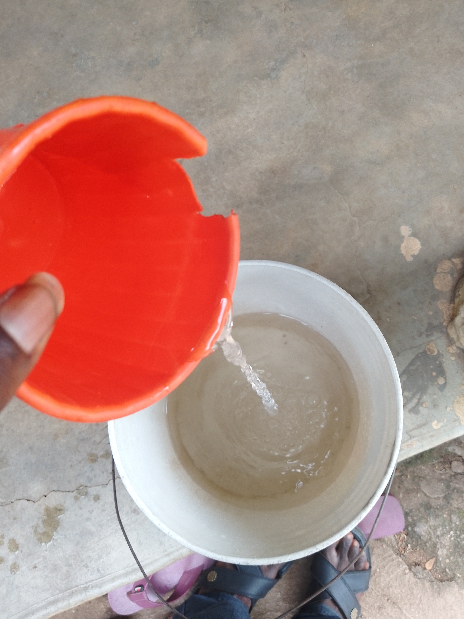 A close-up, overhead shot captures a person's hand pouring water from a cracked, orange plastic cup into a white bucket filled with murky water. The water stream is visible, splashing into the bucket. The ground appears to be a concrete surface with some cracks and dirt marks. In the lower part of the frame, the feet of a person wearing dark sandals and dark trousers are visible, with pink flip-flops underneath. The scene suggests an outdoor setting, possibly in a residential area.