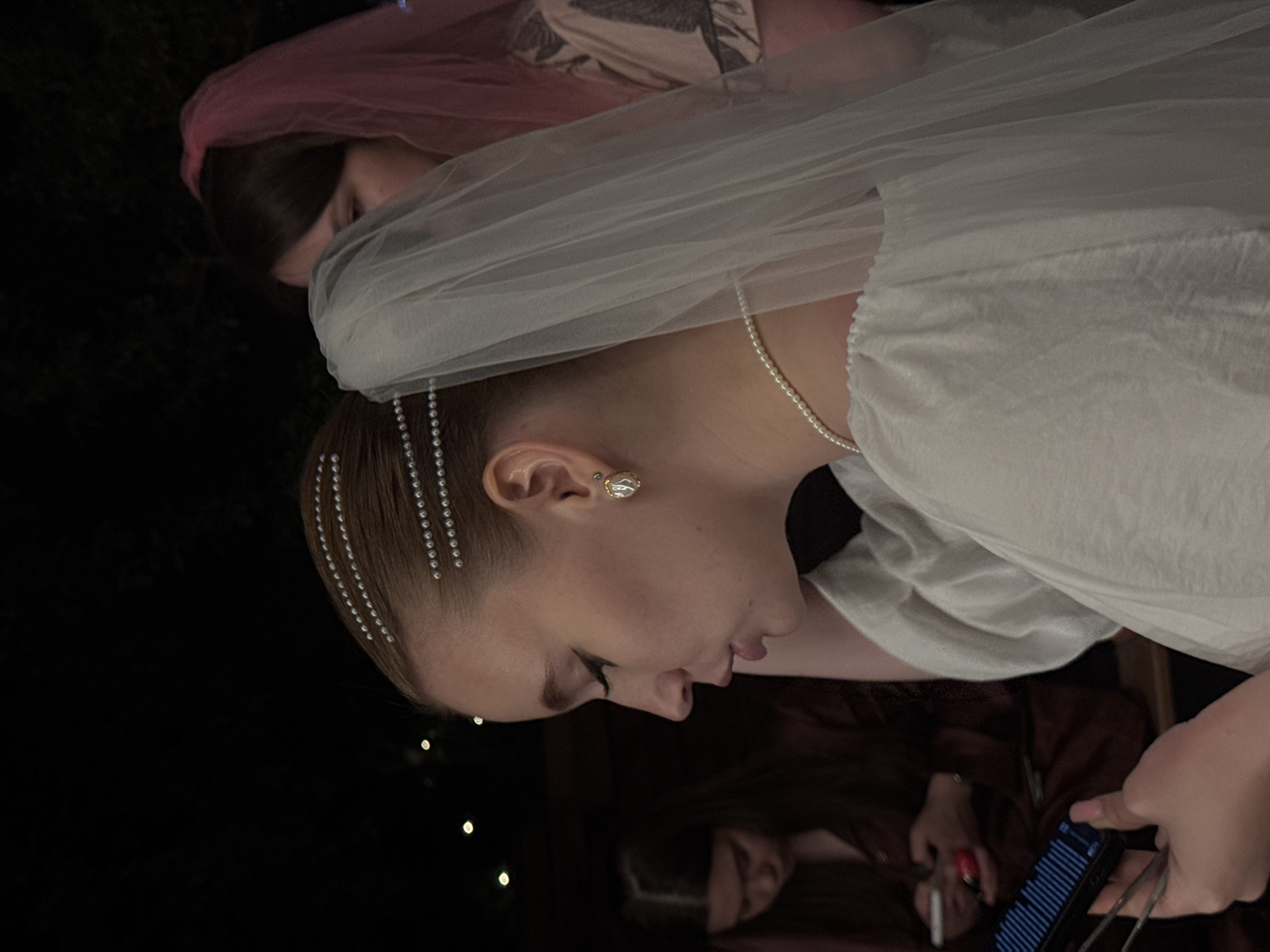 A woman wearing a white veil, a pearl necklace, and a gold earring stands in an outdoor setting. Her hair is decorated with strands of small, clear beads. Two other people are present in the image. One wears a pink veil, and the other is holding a cell phone while sitting in a chair. The scene appears to be dimly lit, with some small lights visible in the background.