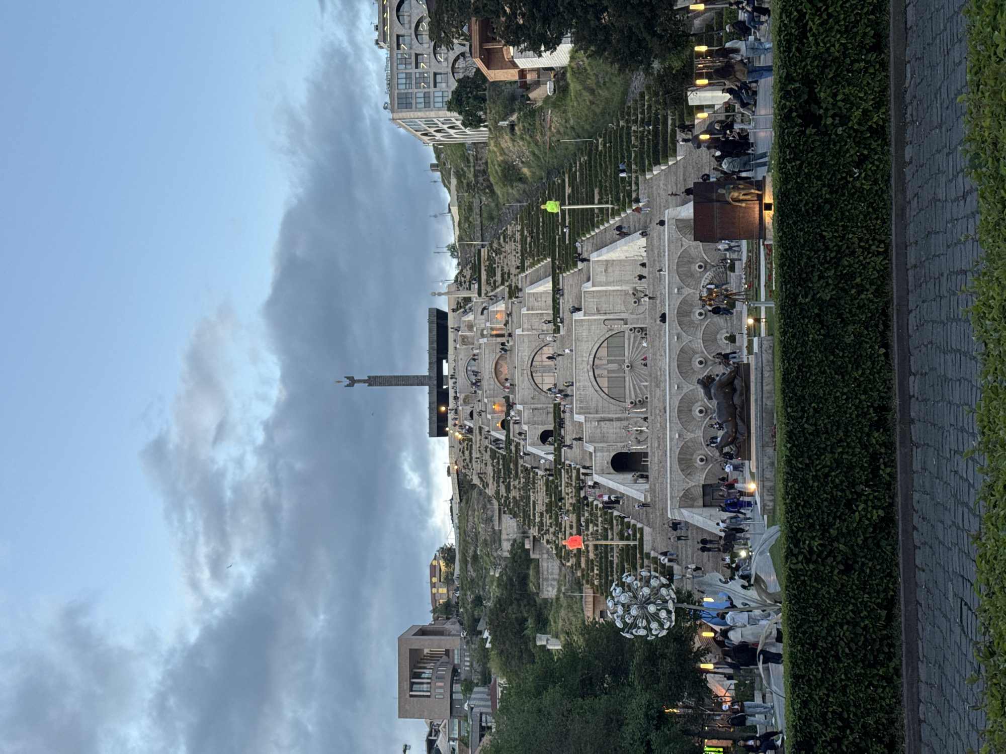 The image depicts the Cascade in Yerevan, Armenia. The Cascade is a monumental stairway, with multiple levels adorned with fountains and sculptures. People are walking along the stairs and viewing the structures. The sky is partly cloudy, suggesting the time of day is dusk. Tall buildings are visible on both sides of the Cascade. The environment is lush with greenery, including hedges and trees.
