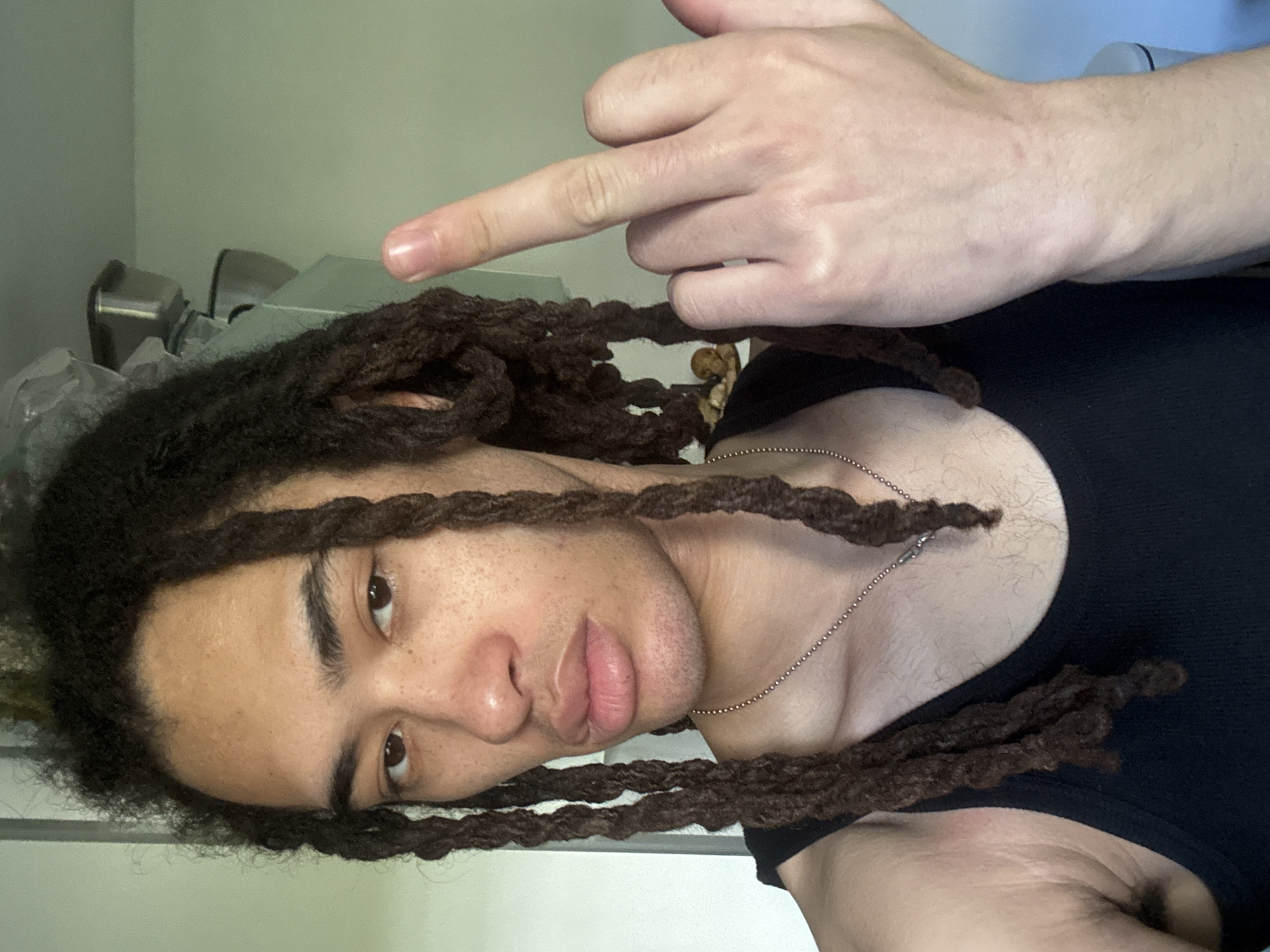 A selfie, rotated ninety degrees counter-clockwise, depicts a person with dark dreadlocks and light-to-medium skin tone, looking directly into the camera. The individual wears a black sleeveless top and a thin, silver ball-chain necklace. Their dreadlocks frame their face, with some strands falling across the chest. The person has dark eyes, distinct eyebrows, and slightly parted lips. There is visible facial hair stubble on the chin and jawline, and some body hair on the chest.
One hand is visible in the upper right portion of the frame, making a gesture with the thumb and index finger extended, while the other fingers are curled. The hand appears to belong to the subject and is positioned to point towards the top-left corner of the image. The person's expression is neutral, suggesting a casual pose.
The background is simple and blurry, indicating an indoor setting. Visible elements include a plain, light-colored wall and some indistinct objects, possibly furniture or fixtures. There is no visible text in the image. The provided context indicates the location is Les Ulis, France, though this is not discernible from the image itself.