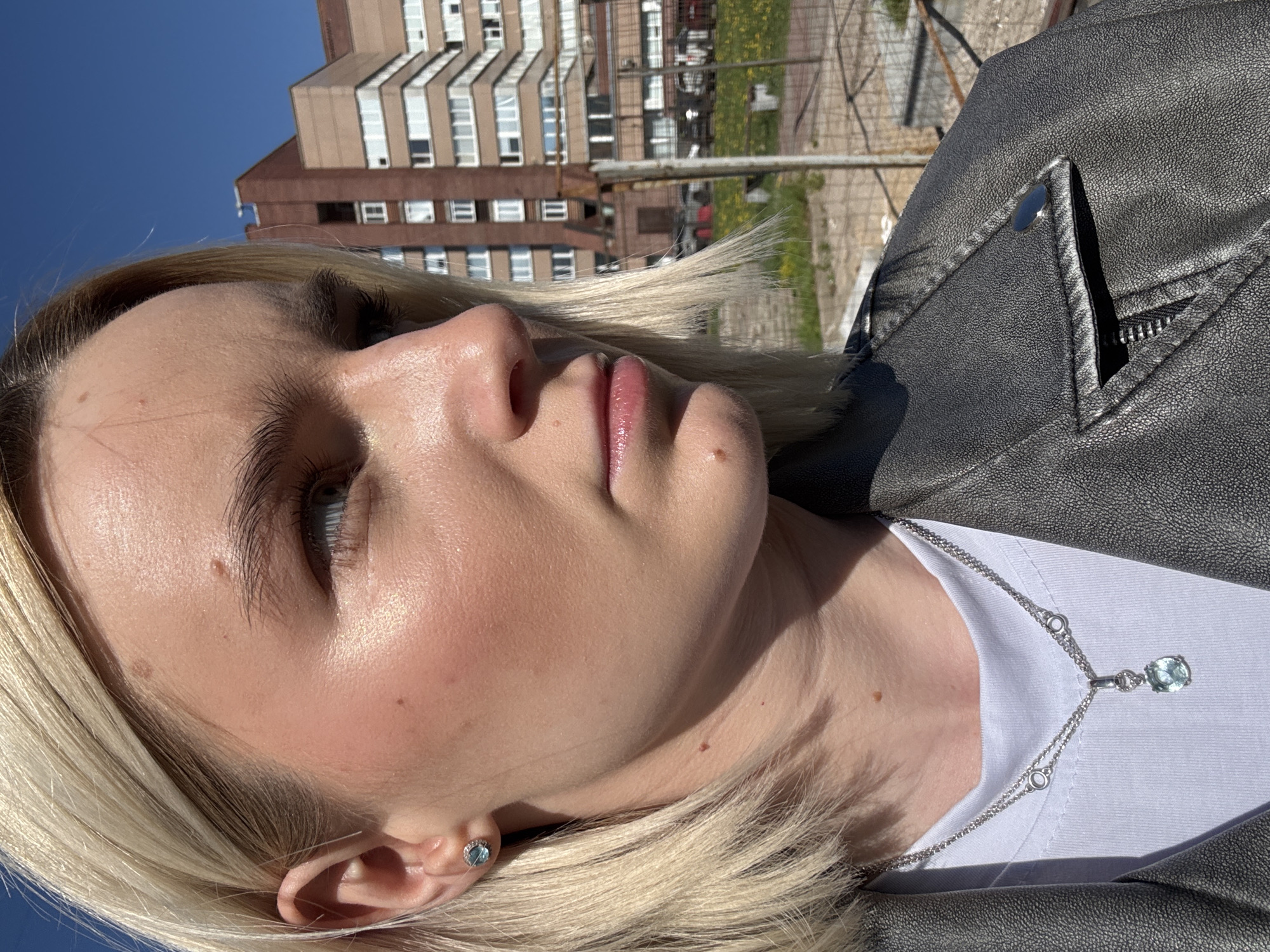 A close-up, rotated portrait captures a fair-skinned blonde woman against an urban backdrop, likely in Saint Petersburg, Russia, given the architectural style of the multi-story building in the background. The woman's face, partially illuminated by sunlight, features fine blonde hair, delicate eyebrows, and light blue eyes. She wears makeup, including blush and lip gloss, and a small, round earring. A silver necklace with a blue stone pendant rests against her white t-shirt. She wears a gray leather jacket. The building behind her is characterized by rows of windows, typical of Soviet-era architecture in Saint Petersburg. A metal fence is visible in the background, along with hints of greenery.