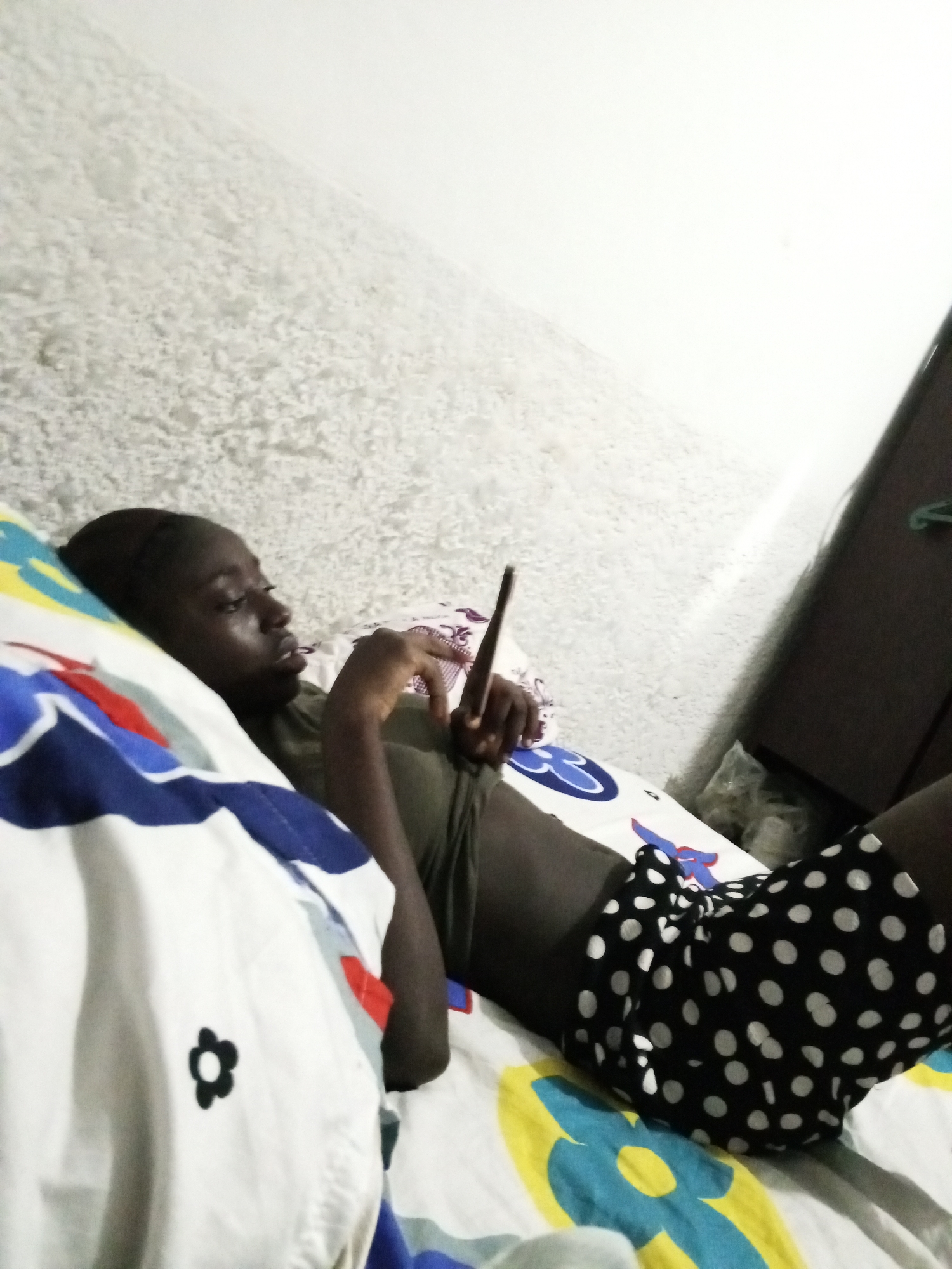 A person is reclining on a bed in a room with a textured white wall. The individual, possibly a young adult, is dark-skinned, wearing an olive-green shirt and black shorts with white polka dots. They are holding a dark, slender object, possibly a phone or electronic device. The bed has multicolored bedding with floral and geometric patterns. A dark-colored wardrobe or cabinet stands in the background near a plastic bag. The location is possibly within a residential area of Abuja, Nigeria, but there is no definitive evidence in the image to confirm that.