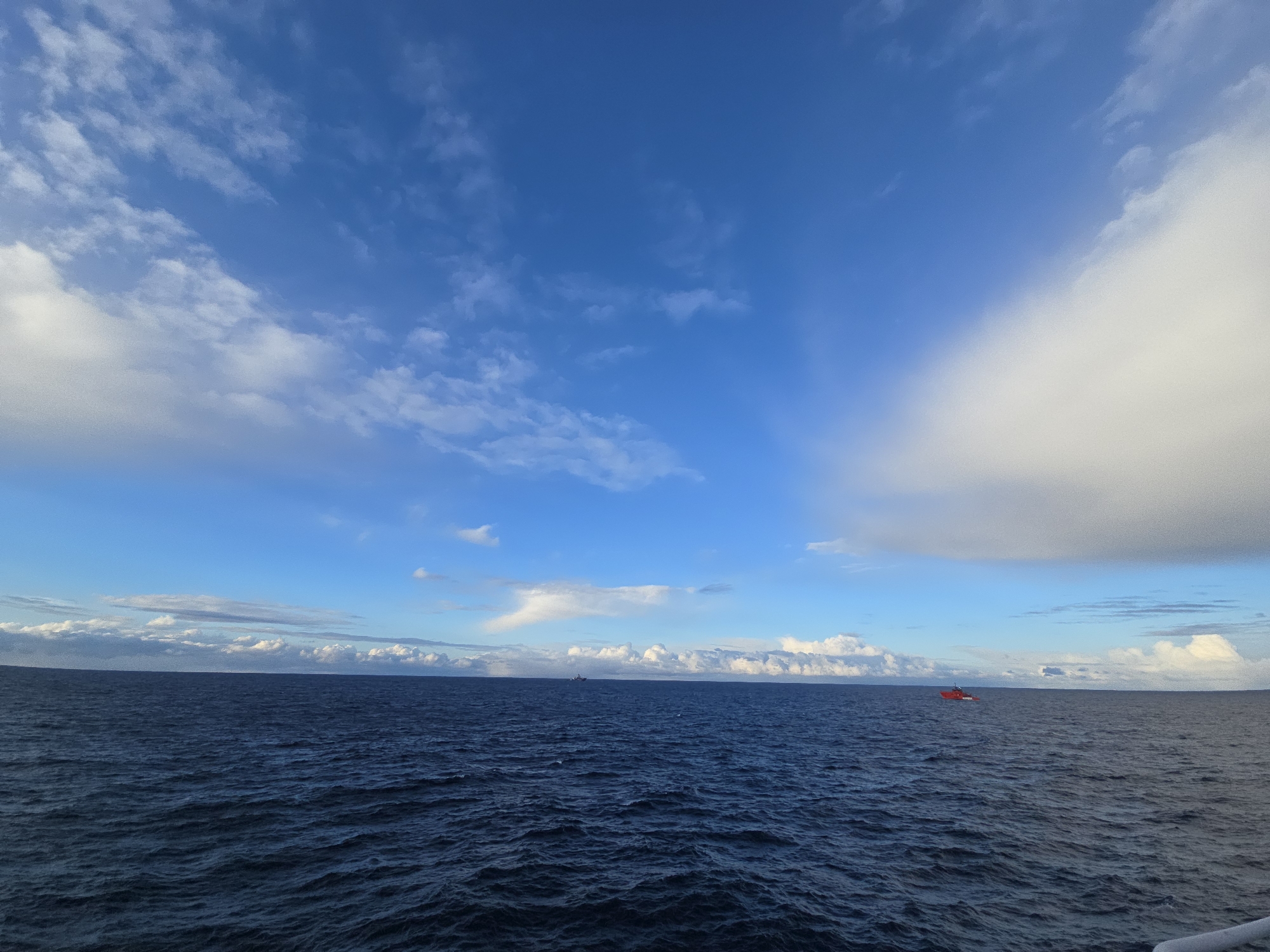 The image depicts a vast expanse of ocean under a partially cloudy sky. The water is dark blue and slightly choppy. A small red boat is visible on the horizon, towards the right side of the image. The sky transitions from a deep blue to a lighter blue, with scattered white clouds throughout. The location is the ocean in the city of Unknown, Unknown.