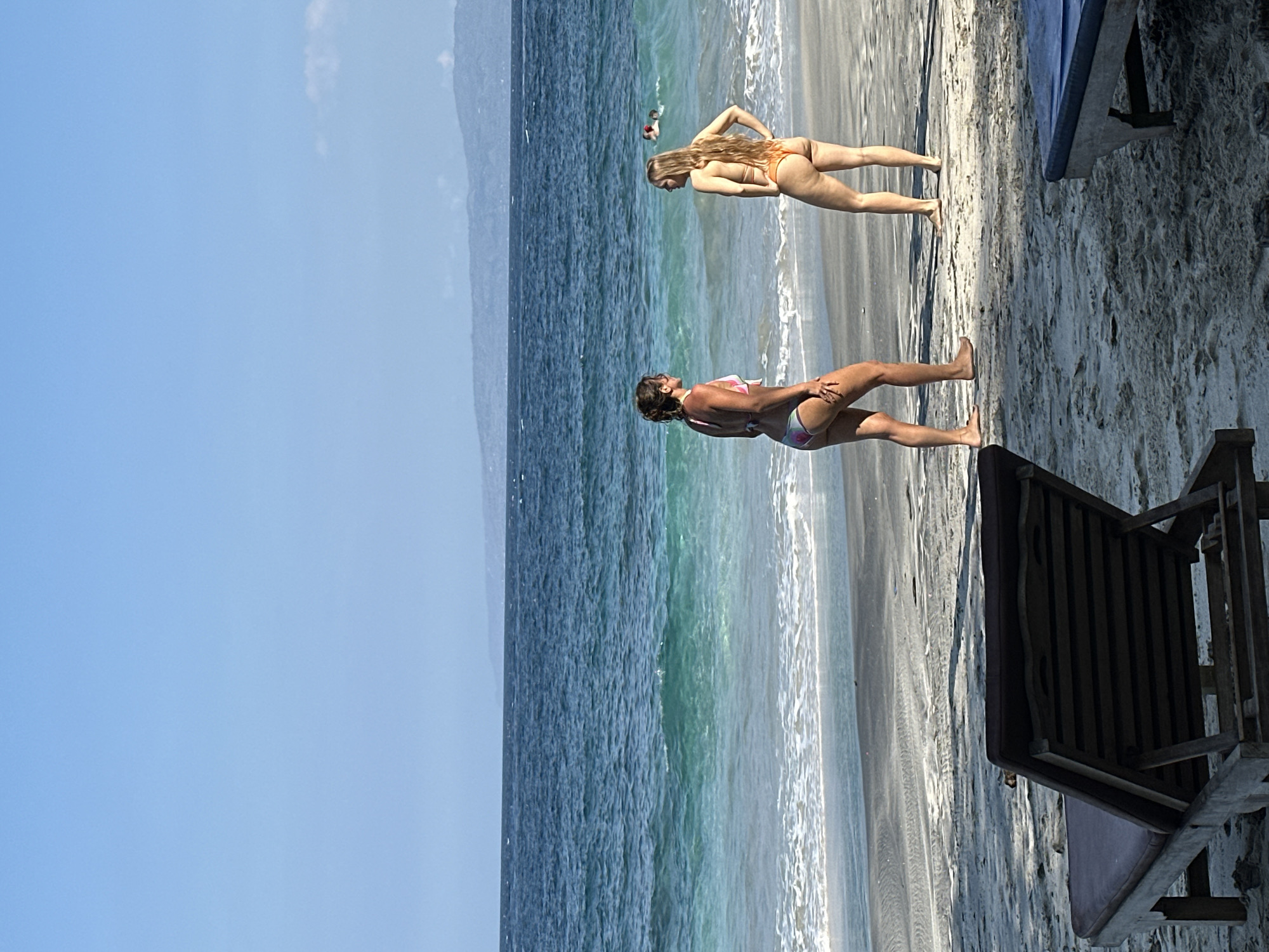 The image depicts a serene beach scene in Manggis, Indonesia, under a clear blue sky. The foreground features a dark-colored lounge chair, partially shadowed, positioned on the sandy shore. In the middle ground, two women are standing on the beach, facing each other and engaged in conversation. The woman on the left wears a multicolored bikini, while the woman on the right is in an orange bikini. The ocean stretches out behind them, with turquoise waters and gentle waves breaking on the shore. Further out at sea, several figures can be seen swimming. The background consists of a hazy, distant coastline with mountains. The overall atmosphere is tranquil and summery.
