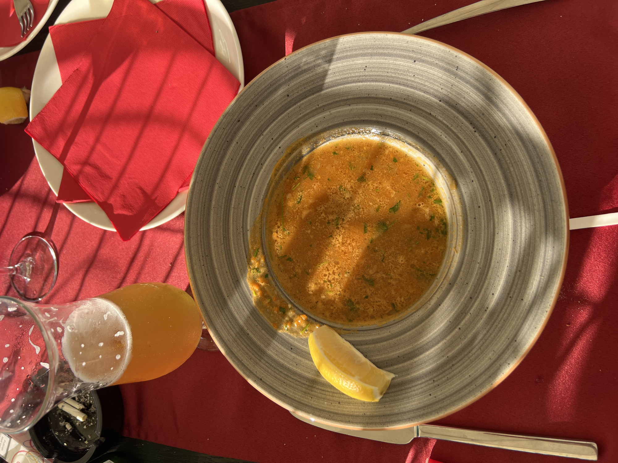 A close-up overhead shot shows a meal set on a red tablecloth in Sveti Stefan, Montenegro. In the foreground, a shallow, grey, speckled bowl contains a vibrant orange soup with visible herbs and vegetables. A wedge of lemon rests on the rim of the bowl. To the left, a fork and knife are placed. In the background, a tall glass of pale golden beer with a frothy head sits next to an ashtray with two cigarette butts. Two red napkins are arranged on a white plate. The scene is illuminated by natural light, casting sharp shadows across the table.