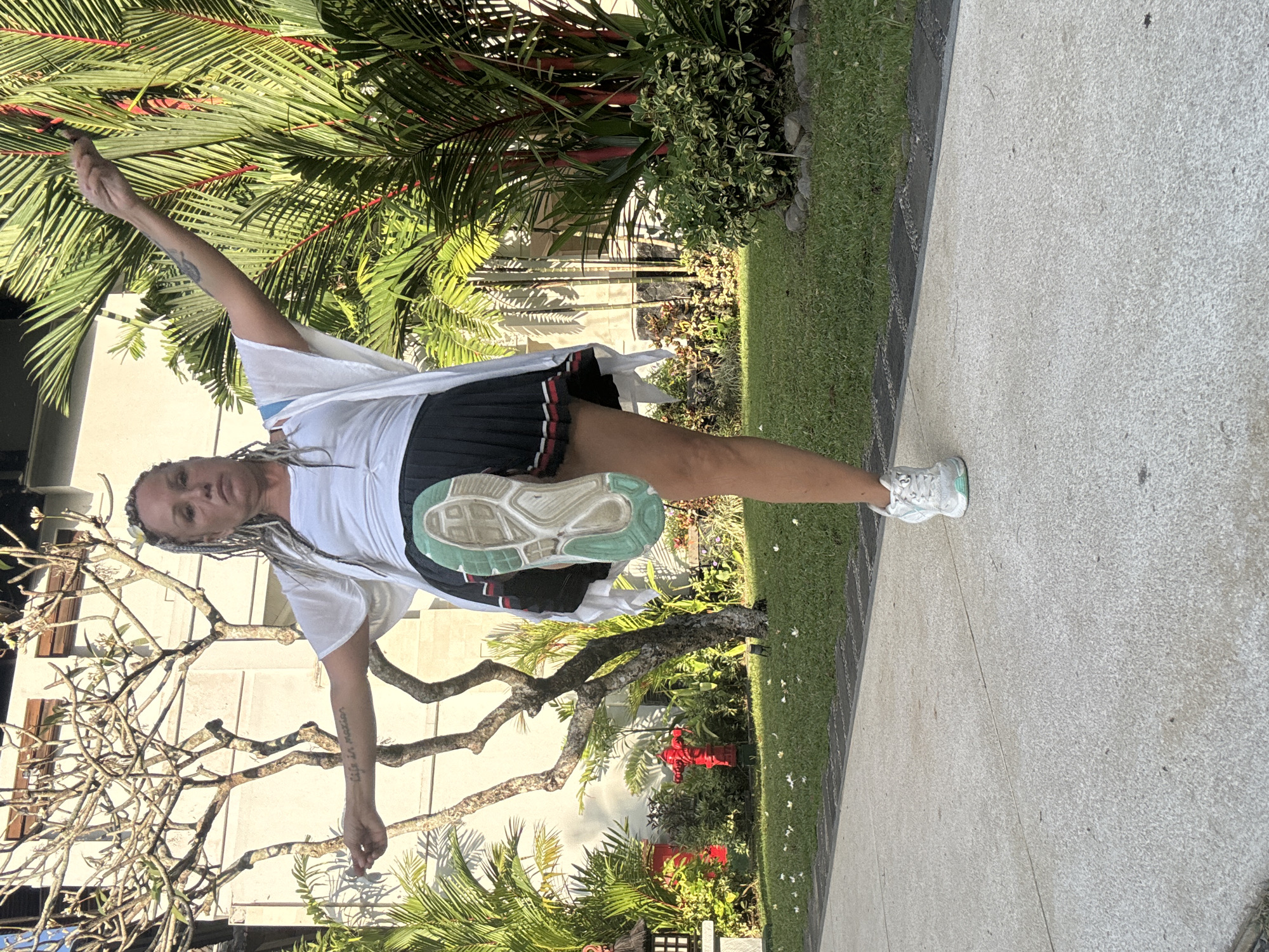 A middle-aged woman with braided blonde hair is performing a standing pose on a concrete walkway in Kuta Selatan, Indonesia. She is wearing a white tank top, a white sheer cardigan, and a black pleated skirt with red and white trim. Her right leg is extended forward, with her right foot lifted and pointing upwards, revealing the sole of her white athletic shoe. Her left leg is supporting her weight. The background features lush green foliage, including palm trees with red stems, a bare-branched tree, and a white building. A red fire hydrant is visible on the grass to the left. The scene is bathed in sunlight, casting shadows on the ground.