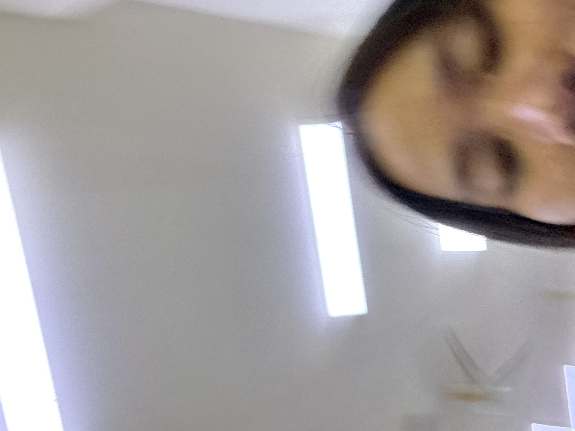 The image is a close-up, out-of-focus shot of a person's face, likely taken from a low angle. The foreground is dominated by the person's forehead, eyes, and nose, with dark hair framing the face. The background consists of a plain, light-colored ceiling with several rectangular fluorescent light fixtures. One prominent light fixture is directly above and slightly to the left of the person's head, emitting a bright white light. The overall impression is of an indoor environment, possibly an office or classroom, with a sterile, well-lit atmosphere. The location in East Jakarta, Indonesia, is not discernible from the visual information provided in the image itself.