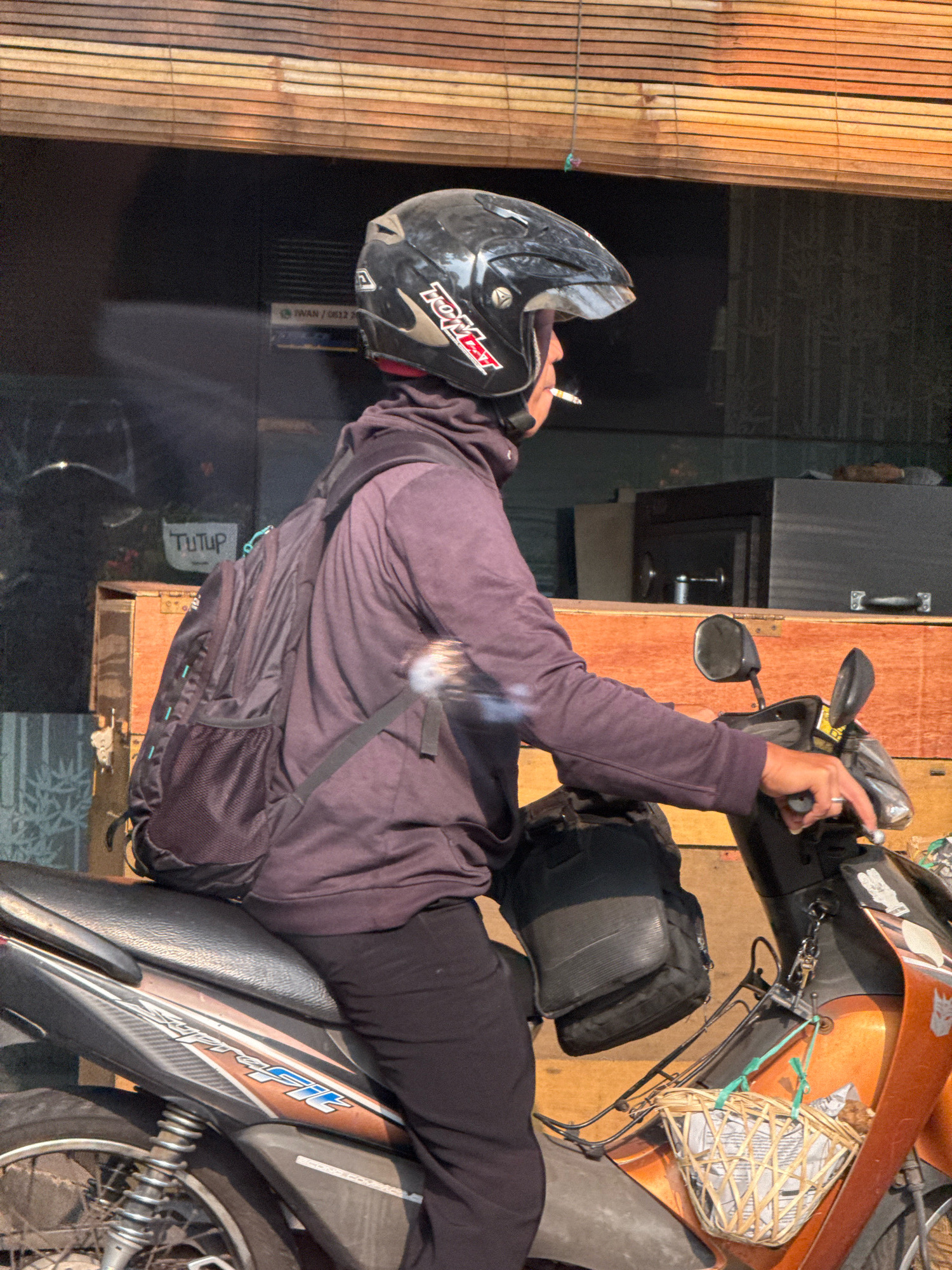 A person wearing a black helmet and a purple hoodie is riding a motorcycle in Surabaya, Indonesia. The person is smoking a cigarette, and there is smoke visible in the image. The motorcycle is orange and black, and it has a basket attached to the front. The background of the image is a building with a wooden blind. There is a sign that says "TUTUP" which means "CLOSED" in Indonesian. The overall lighting suggests it is daytime.