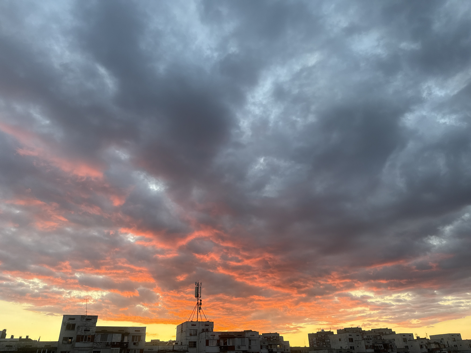 A low-angle shot depicts a dramatic sunset over a city skyline. The sky is filled with dark, grey storm clouds above, juxtaposed with a band of intense orange and yellow light near the horizon, illuminating the clouds from below. A telecommunications tower stands atop one of the buildings, silhouetted against the colorful sky. The buildings are low to mid-rise, typical of urban residential architecture, and appear to be closely spaced.
While the image itself does not confirm the location, it visually aligns with the general architectural style and urban landscape found in parts of Sofia, Bulgaria. The buildings suggest a district with Soviet-era architecture, common in Sofia.