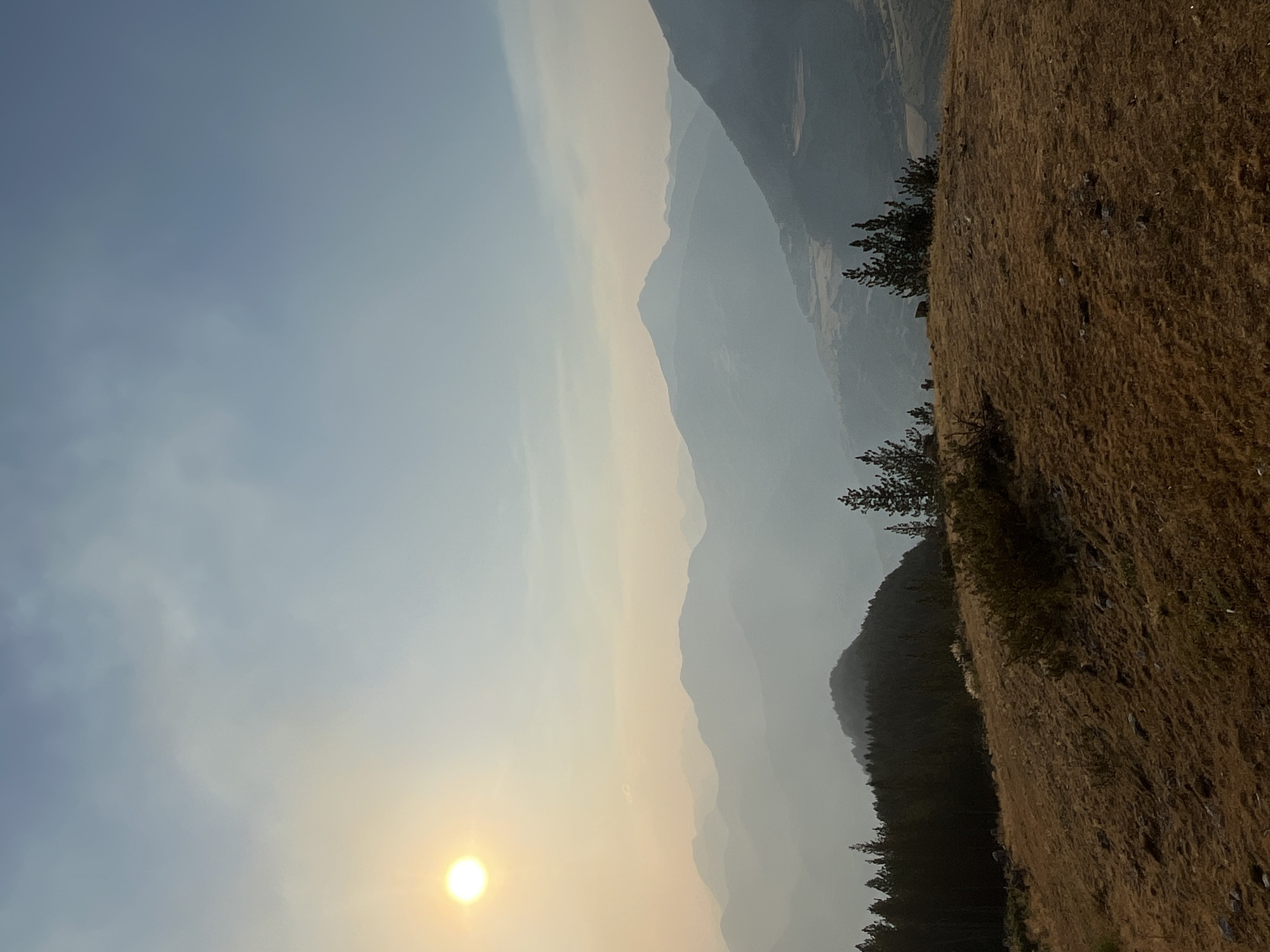 The image depicts a hazy mountain landscape at sunset. The sun, a bright, hazy orb, is positioned in the upper left quadrant, casting a soft glow. In the foreground, a dry, grassy hilltop slopes downward, sparsely dotted with rocks and low-lying scrub brush. A cluster of dark green pine trees stands on the left side of the frame, bordering the crest of the hill. Beyond the foreground, a series of mountainous ridges recede into the distance, their forms softened by atmospheric haze and the fading light. The sky transitions from a pale, hazy yellow near the sun to a clear, light blue at the top. There are no visible people or animals in the scene, and the environment appears to be a natural, undeveloped mountainous region. The location is identified as San Juan de Plan, Spain.