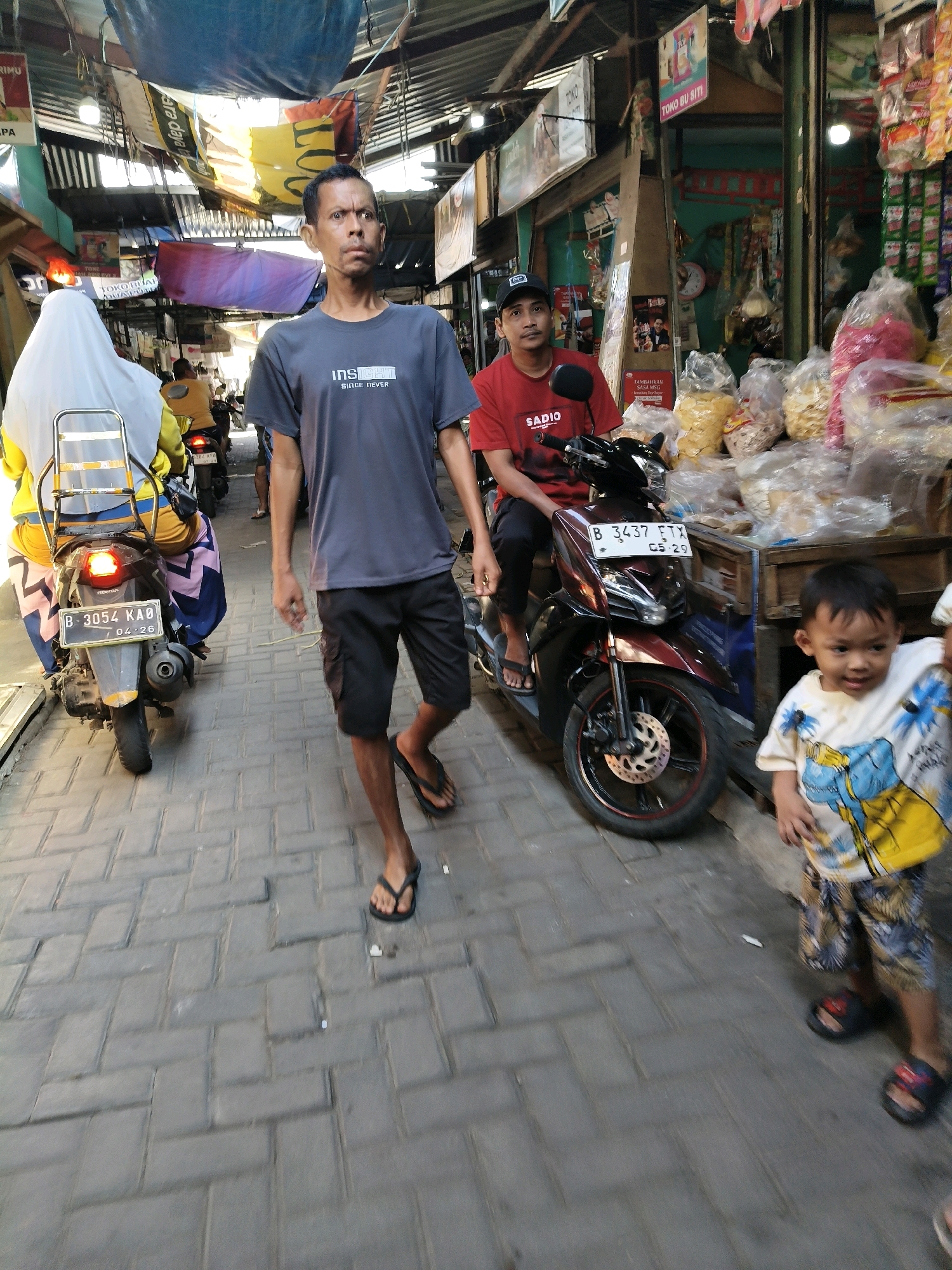 The image shows a street scene in Setiamekar, Indonesia. A man in a dark grey t-shirt and black shorts walks towards the camera. To his left, a woman wearing a white hijab and yellow top rides a scooter. In the background, a man in a red t-shirt sits on a motorcycle next to a stall selling bagged snacks. A young boy in a colorful t-shirt and patterned shorts stands in the foreground to the right of the frame. The street is paved with interlocking bricks, and the area is lined with shops and stalls, suggesting a marketplace environment. Overhead, tarps and signs are strung across the street, providing shade.