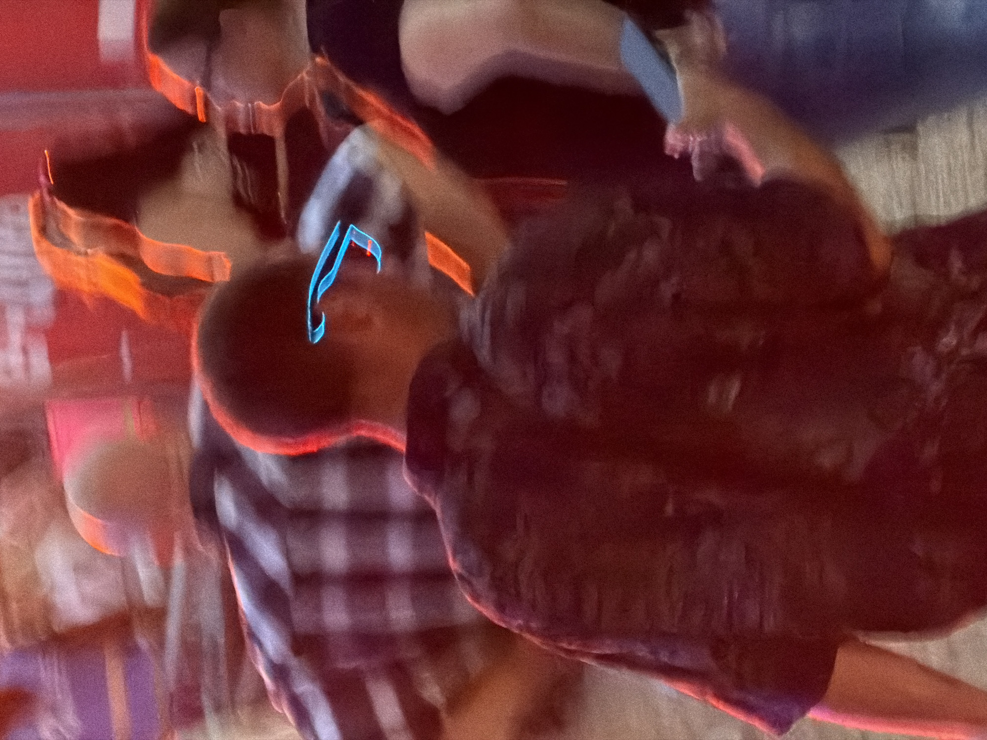 The image depicts a blurry, nighttime scene with several individuals. A person in the foreground, wearing a dark, patterned shirt, is partially visible from the back. To their left, another person is seen wearing a plaid shirt. In the background, a person with blonde hair is illuminated by orange and red lights, and another individual in the right side of the frame is wearing glasses. A neon blue object, possibly part of sunglasses or an accessory, is prominently displayed in the center of the image. The overall setting appears to be an outdoor event or gathering, possibly in Echternach, Luxembourg, given the context. The low light and motion blur suggest a lively atmosphere, possibly a festival or concert.