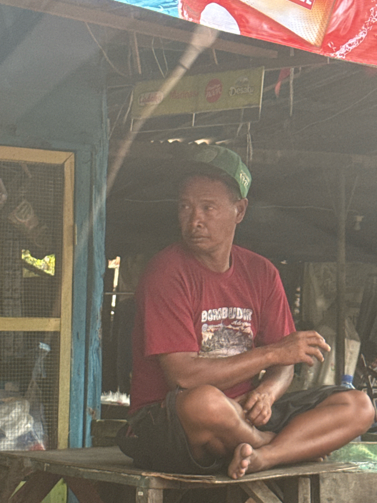 A man wearing a red t-shirt with "BOROBUDIR" printed on it, and a green cap, is sitting cross-legged on a wooden surface. He is looking to his left. The man appears to be in an outdoor setting, possibly a market stall or a roadside establishment. In the background, there is a blue wooden structure with a wire mesh door. A red and white banner is visible overhead. The scene suggests a casual, everyday moment in what could be a commercial area in Surabaya, Indonesia.