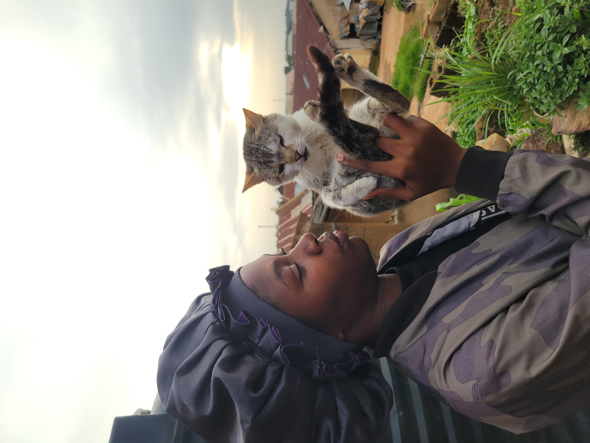 A person wearing a dark satin bonnet and a camouflage bomber jacket holds a grey and white cat in their arms, facing towards the right of the frame. The cat is looking slightly down and to the left, with its paws extended. The background depicts a residential area in Jos, Nigeria, characterized by corrugated iron roofs and scattered vegetation. The sky is overcast with diffused sunlight, suggesting it might be early morning or late afternoon. The person's lips are slightly pursed, as if about to kiss the cat. The image is captured outdoors, with a slightly elevated perspective looking down on the person and cat.