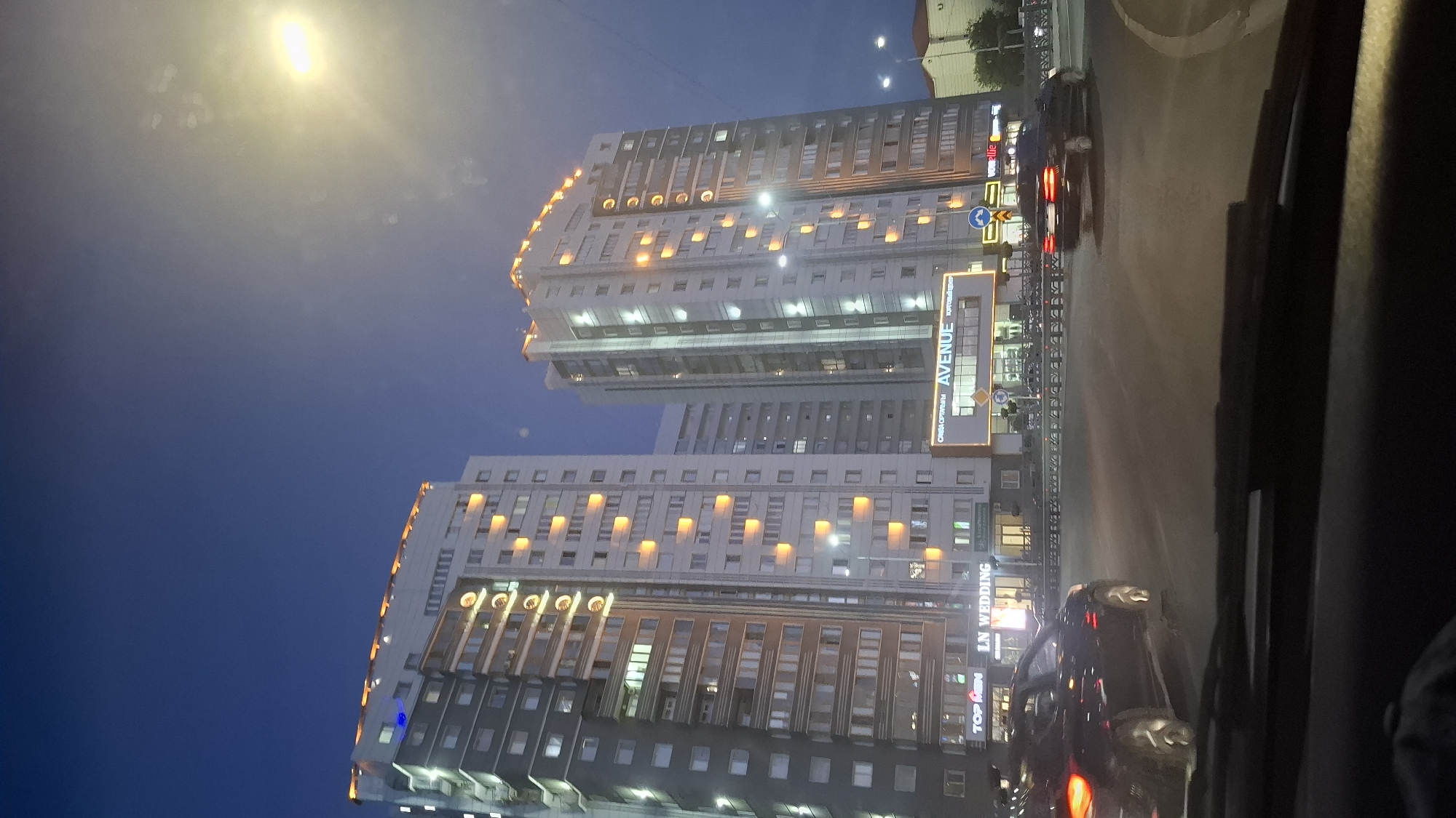 The image shows a street scene at dusk in Shymkent, Kazakhstan, focusing on modern high-rise buildings illuminated by interior and exterior lights. In the foreground, the dashboard of a vehicle is visible, suggesting the photo was taken from inside a car. Two prominent apartment buildings dominate the skyline. The building on the left is taller and wider, featuring a facade with numerous windows and vertical light strips. The building on the right is also a high-rise, slightly set back, with similar architectural features and lighting.
At the base of the buildings, a commercial strip is visible with illuminated signs. Notable signs include "TOP MEN," "LN WEDDING," and "AVENUE ТОРГОВЫЙ ЦЕНТР" (AVENUE SHOPPING CENTER). Several cars are on the road, with a black car visible in the middle distance turning. Traffic lights and streetlights are also present. The sky is a deep blue, transitioning to lighter shades near the horizon where a bright streetlamp is prominently visible, casting a strong yellow glow and creating lens flare. The overall impression is a well-lit urban environment at the end of the day.