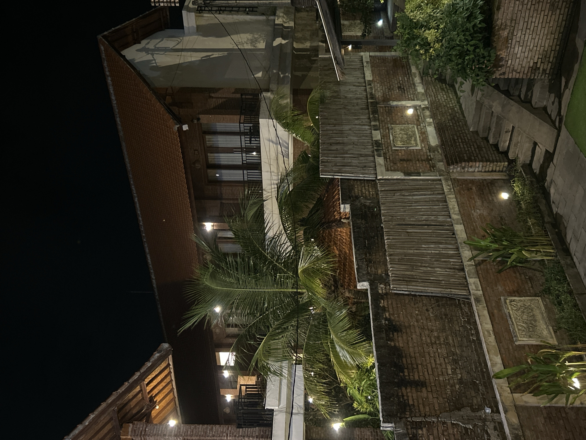 This nighttime exterior photograph captures a multi-story brick building in North Kuta, Indonesia, likely a hotel or guesthouse. The building features red-tiled roofs and balconies with black railings. A prominent palm tree with large fronds is situated in the foreground, partially obscuring the structure. The scene is illuminated by warm-toned lights, including string lights and wall-mounted fixtures, creating a soft glow. The architecture incorporates exposed brickwork and bamboo elements. A set of concrete stairs leads upwards to the right side of the frame. No people are visible in the image. The overall impression is one of a tropical, illuminated evening ambiance.