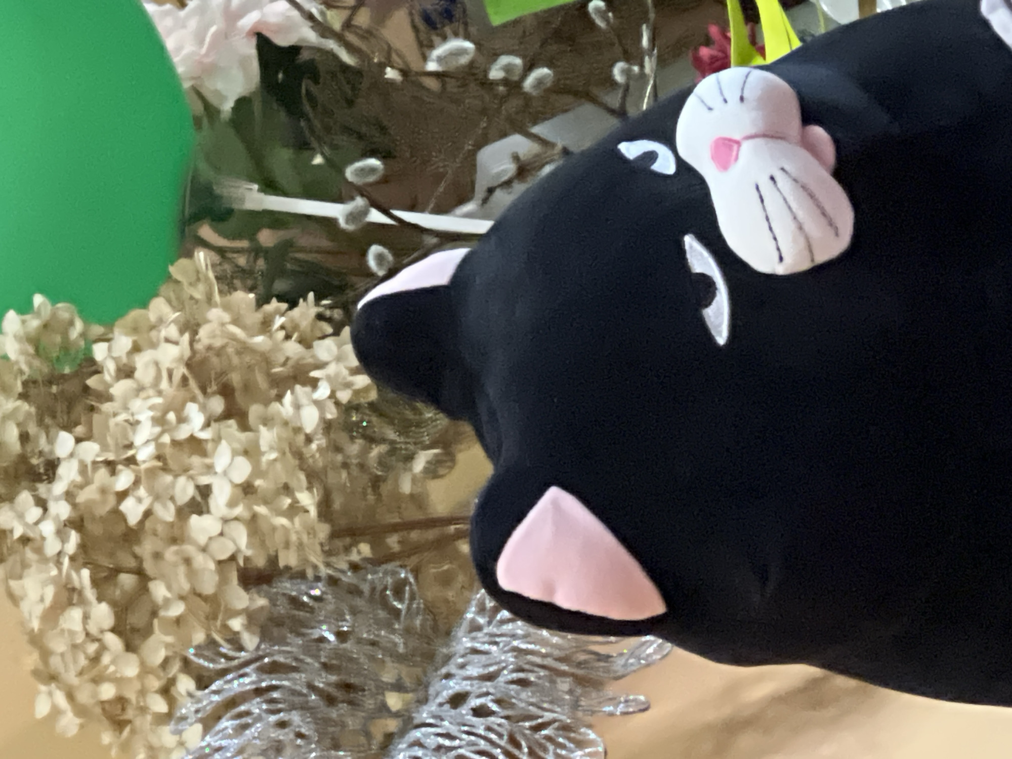 The image shows the back of a black stuffed cat with pink ears. The cat's face is partially visible, featuring white embroidered eyes and a pink nose with white whiskers. In the background, there is a bouquet of dried flowers, a silver tinsel decoration, and a bright green balloon. The scene is likely indoors, possibly in a home or event space. No specific location in Minsk, Belarus is identifiable from the image.
