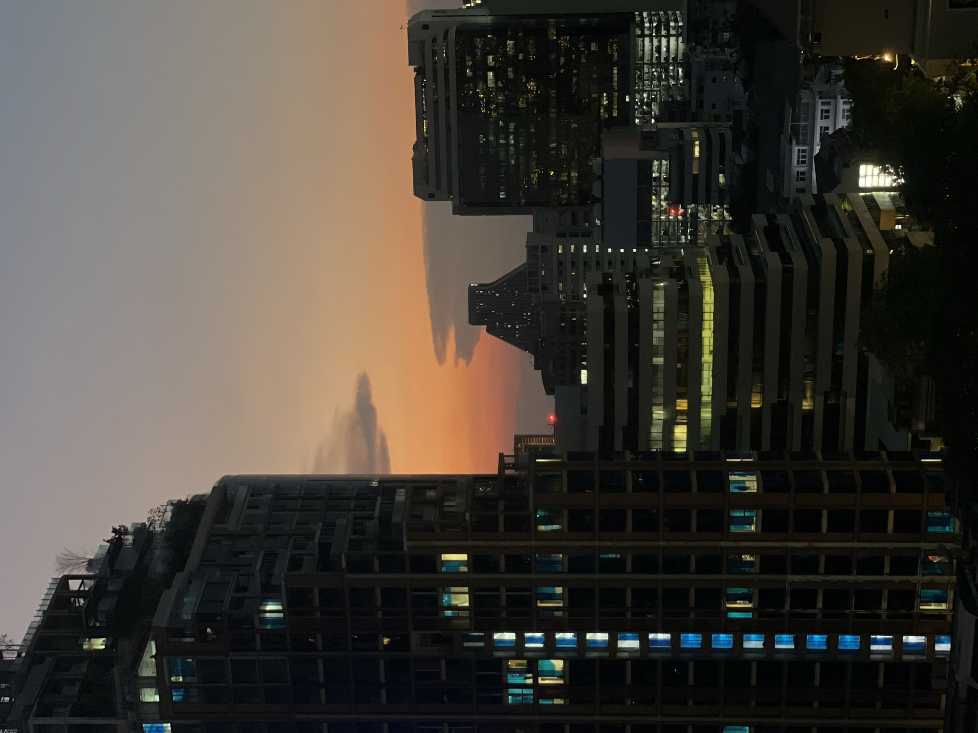 The image shows an evening cityscape in Bangkok, Thailand. Several tall buildings dominate the view, with many windows illuminated. The sky transitions from a dark gray to orange, suggesting either a sunset or sunrise. There are no people visible in the shot. The buildings appear to be modern structures with a mix of residential and commercial usage. The environment is urban, emphasizing the dense city environment.