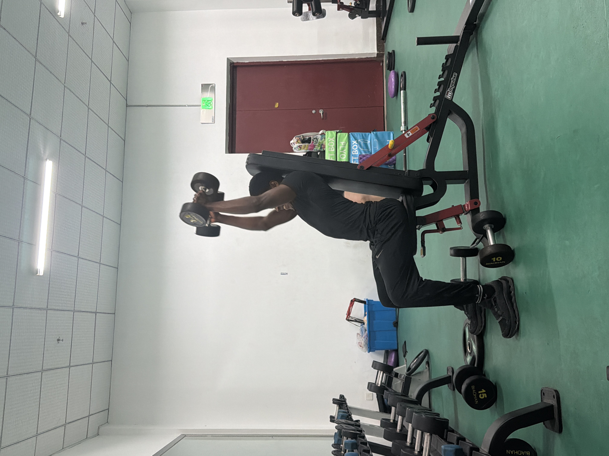 A person is in a gym, performing an exercise while lying prone on a weight bench. The person is holding dumbbells. The gym has green flooring and white walls. Other weights and equipment are visible in the background. The location is likely in Jiangning District, China due to text on the weights reading "BIAOHAN".