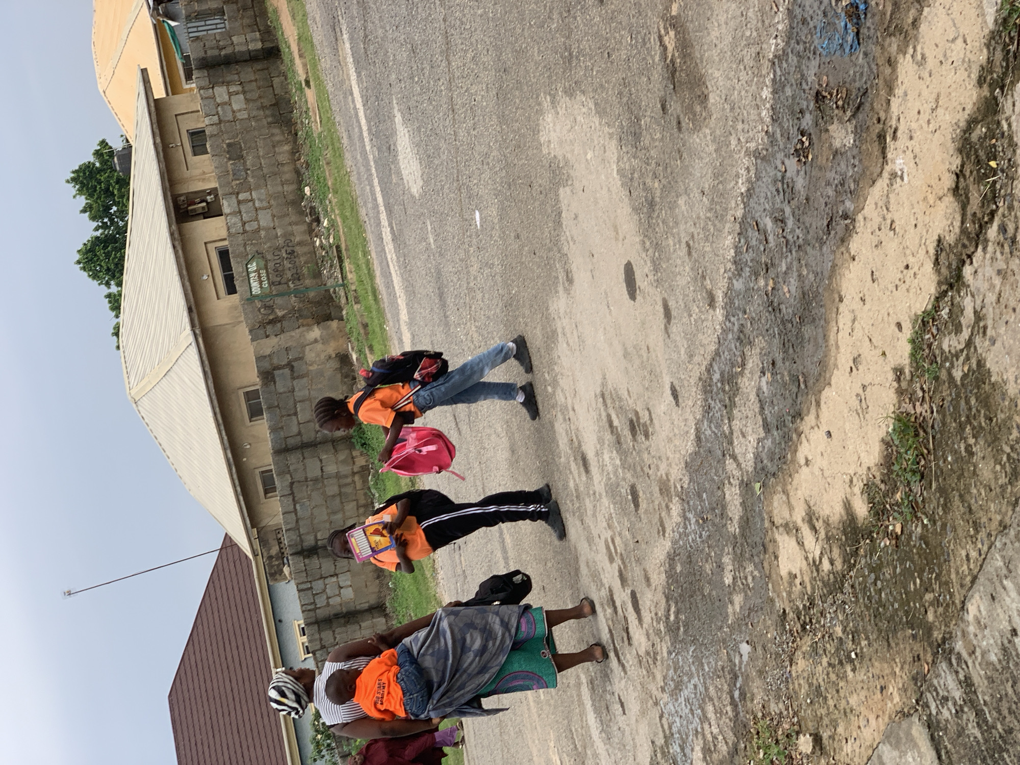 In Abuja, Nigeria, a woman in a patterned headscarf and a traditional wrapper carries a young child on her back. She stands on a paved road next to two school-aged children. One child, wearing an orange shirt and black pants, holds a book. The other, in an orange shirt and blue jeans, carries a pink backpack. Behind them, a wall made of grey bricks separates the road from a residential area. Houses with beige and light yellow roofs are visible, and trees line the background. The sky is overcast.