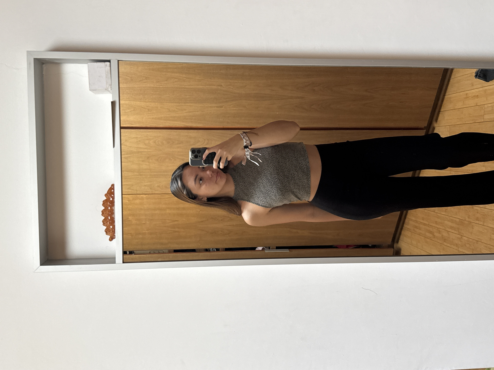 A young woman is taking a selfie in a mirror. She is wearing a gray tank top and black leggings. The mirror is framed in a gray metal frame and reflects a wooden cabinet and a light-colored wooden floor. The background also shows a white wall. Based on available data, a location in San Bernardino Tlaxcalancingo, Mexico cannot be determined.